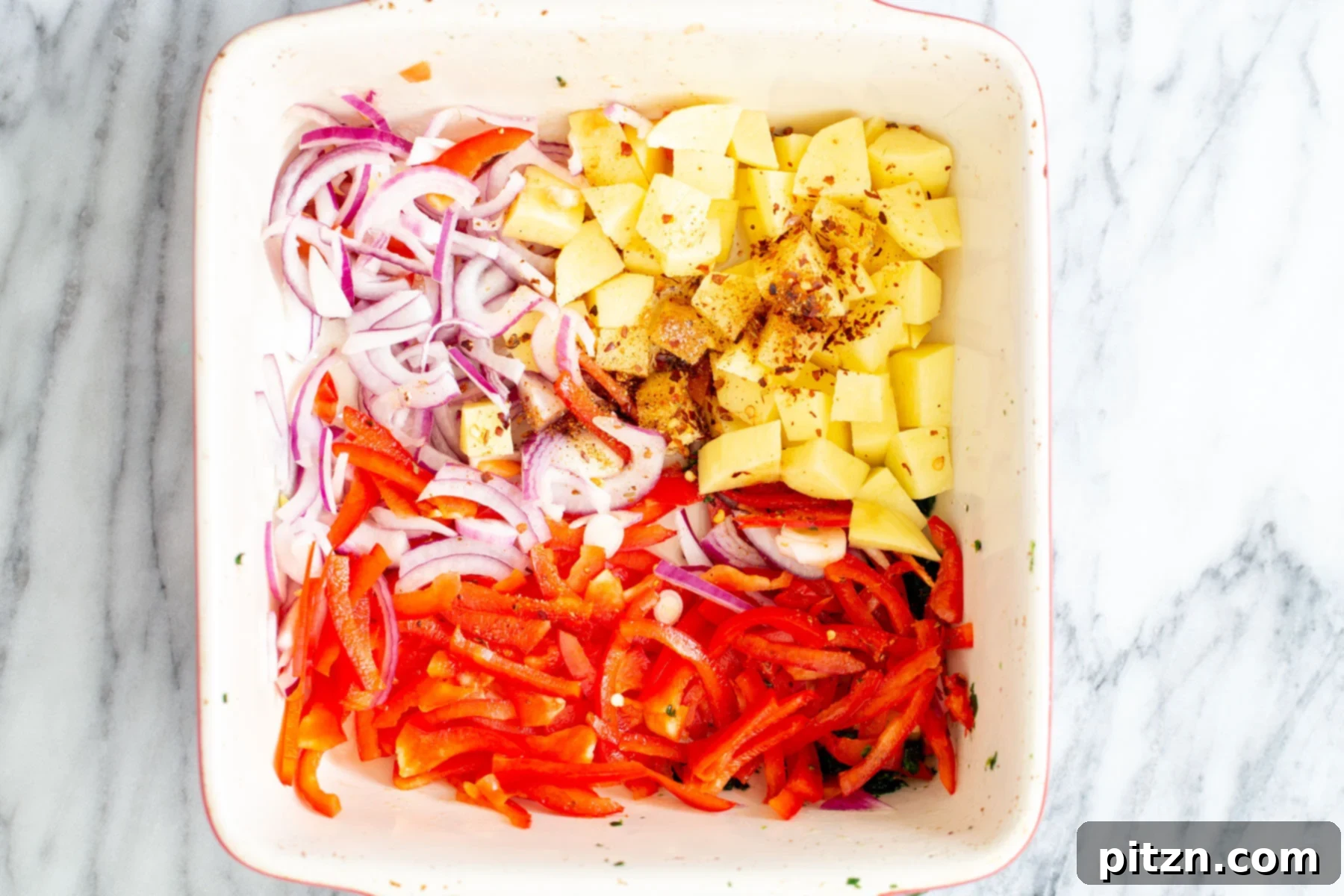 Sliced onion and bell pepper, along with cubed potatoes, arranged in a white casserole dish before roasting.