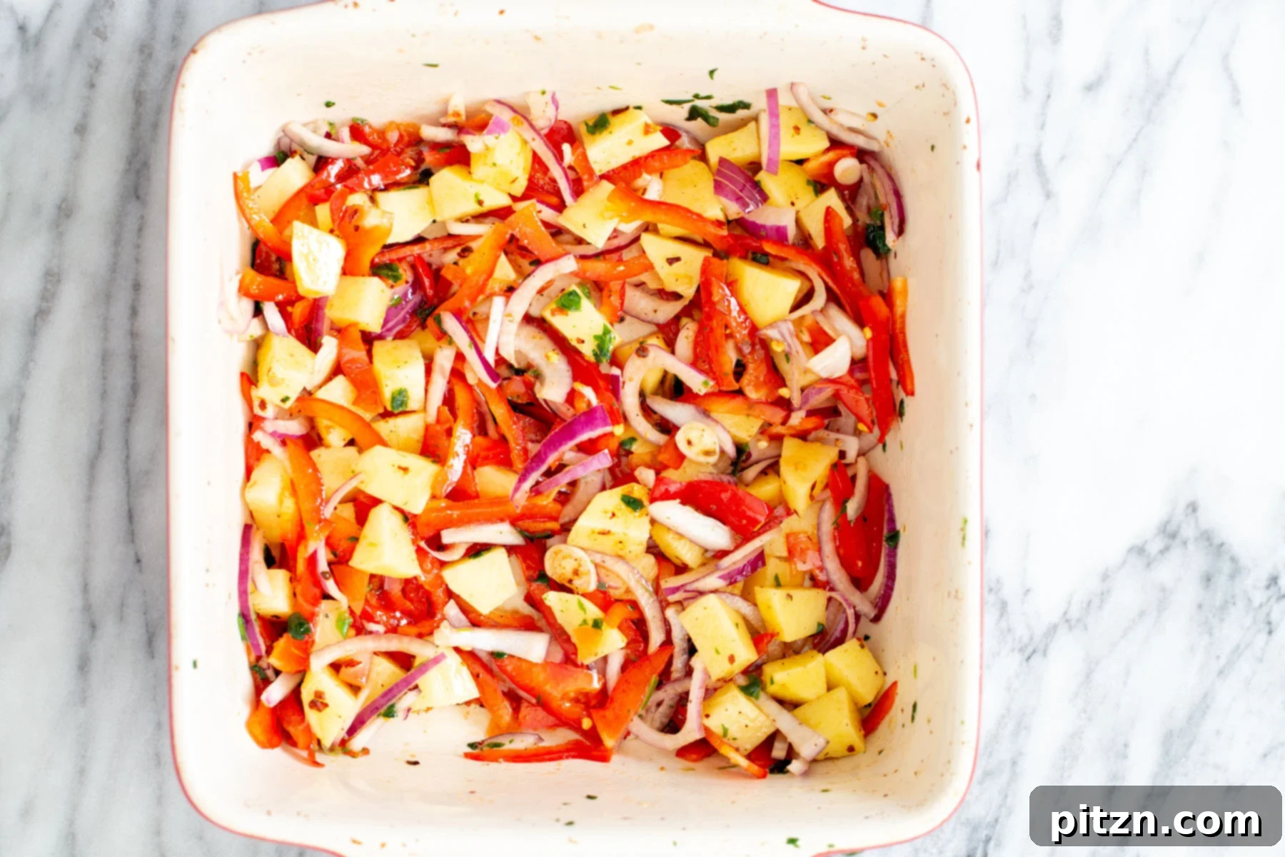 Cubed potatoes, sliced onions, and bell peppers are mixed with seasoning and oil in a white baking dish, ready for roasting.