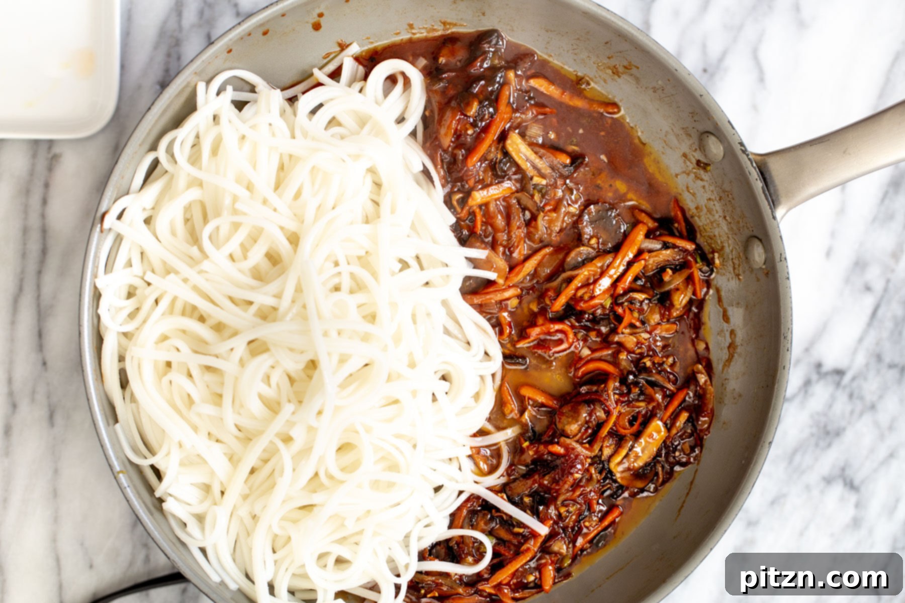 A close-up of perfectly cooked rice noodles and vibrant vegetables coated in a rich Korean stir-fry sauce, all mixed together in a saucepan.