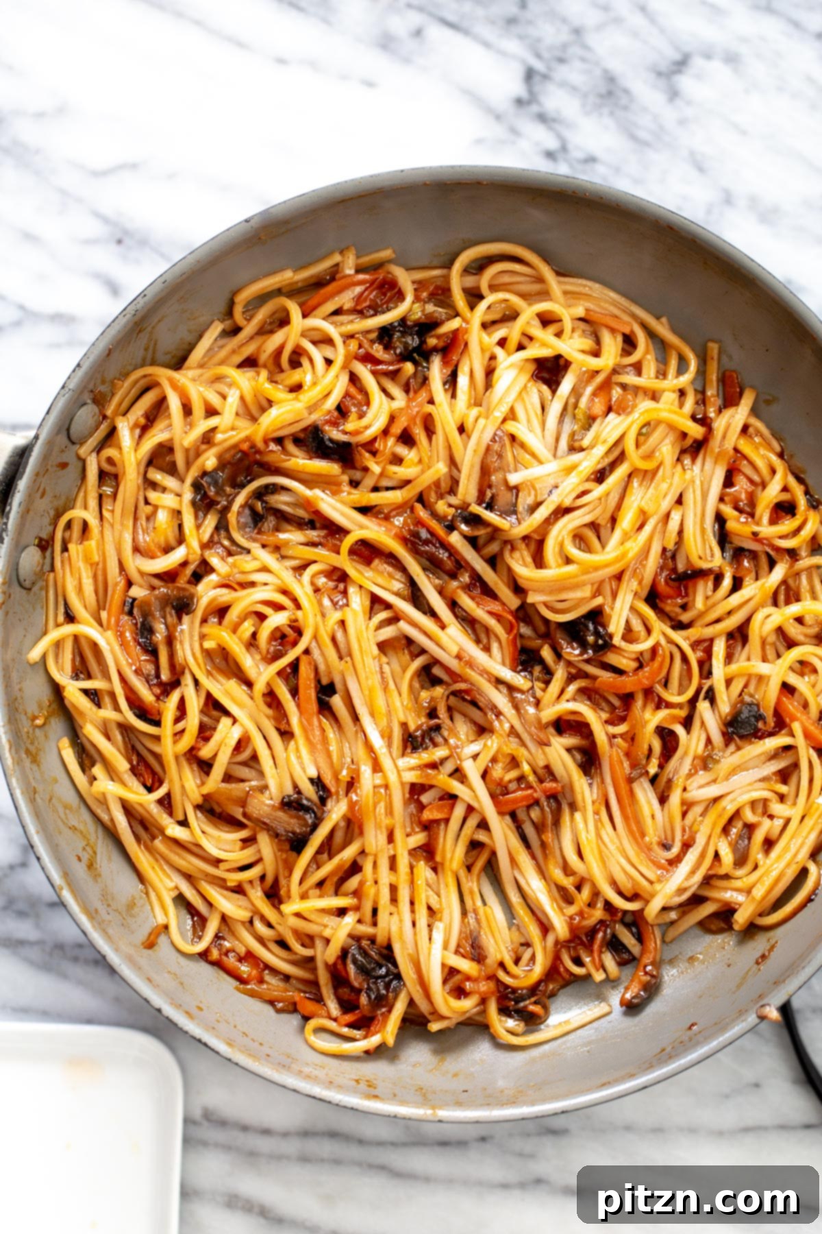 A wide shot of vegan Korean gochujang noodles, glistening with sauce and mixed with vegetables, resting in a large saucepan after cooking.