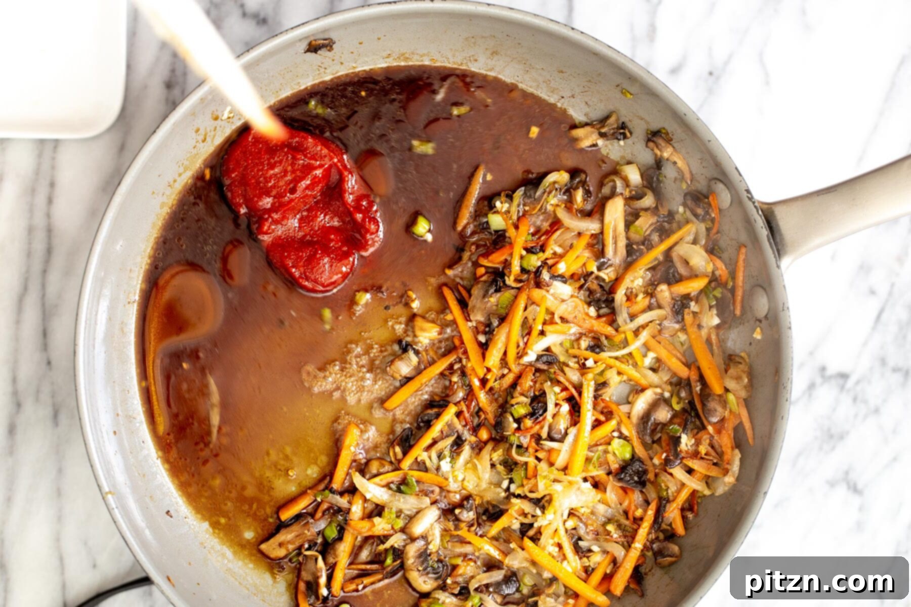 A hand adding gochujang sauce ingredients to sautéed vegetables in a saucepan, showcasing the creation of the vibrant Korean stir-fry sauce.