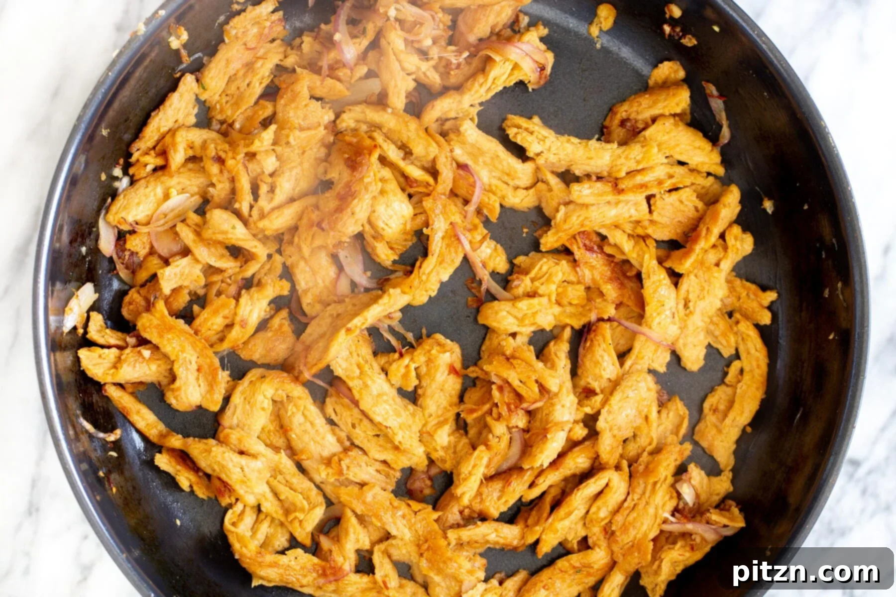 Close-up of marinated soy curls searing in a frying pan alongside garlic and onions, developing golden edges