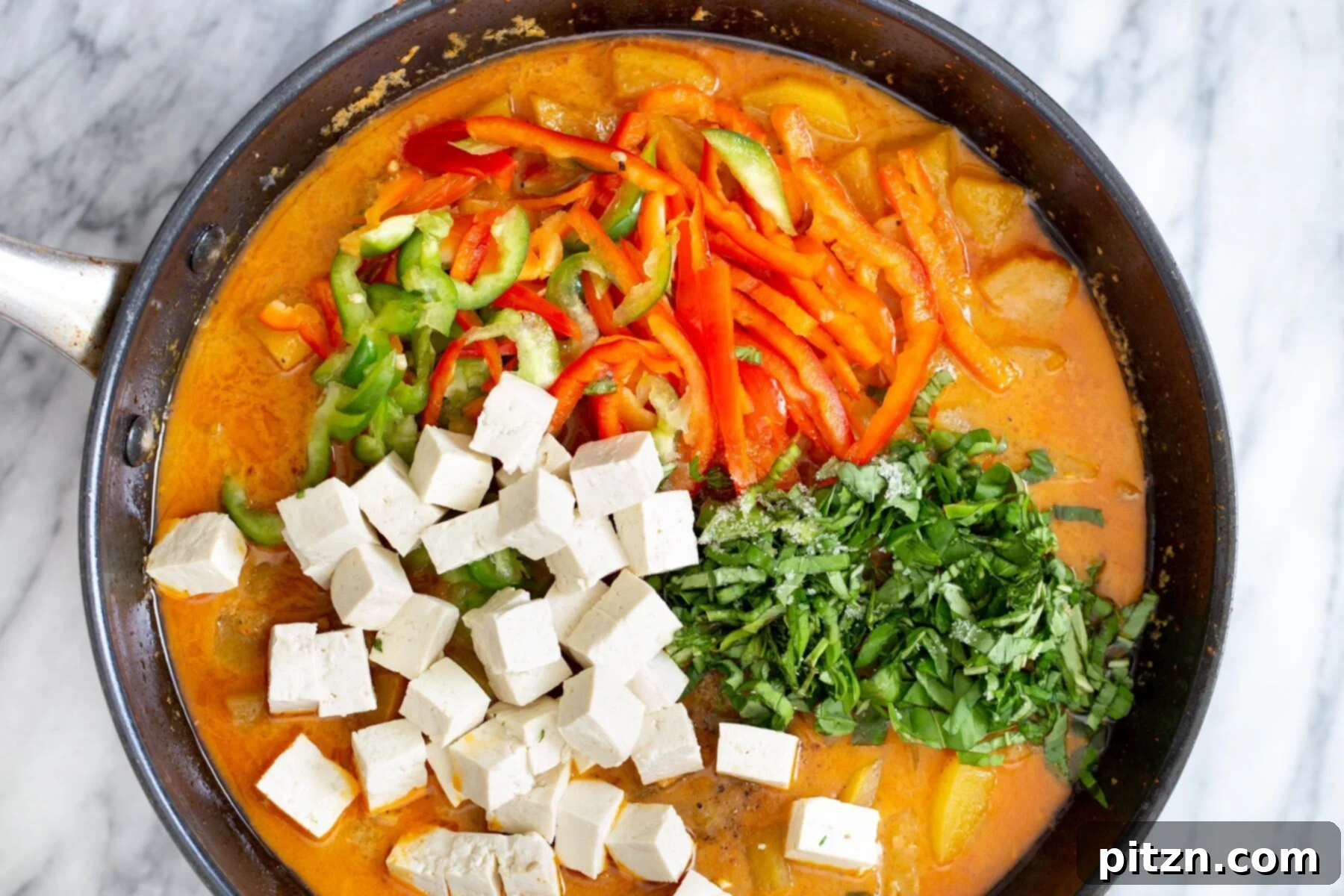 Cubed tofu, colorful bell peppers, and fresh Thai basil being added to the simmering pumpkin coconut broth in a saucepan, enhancing color and flavor.