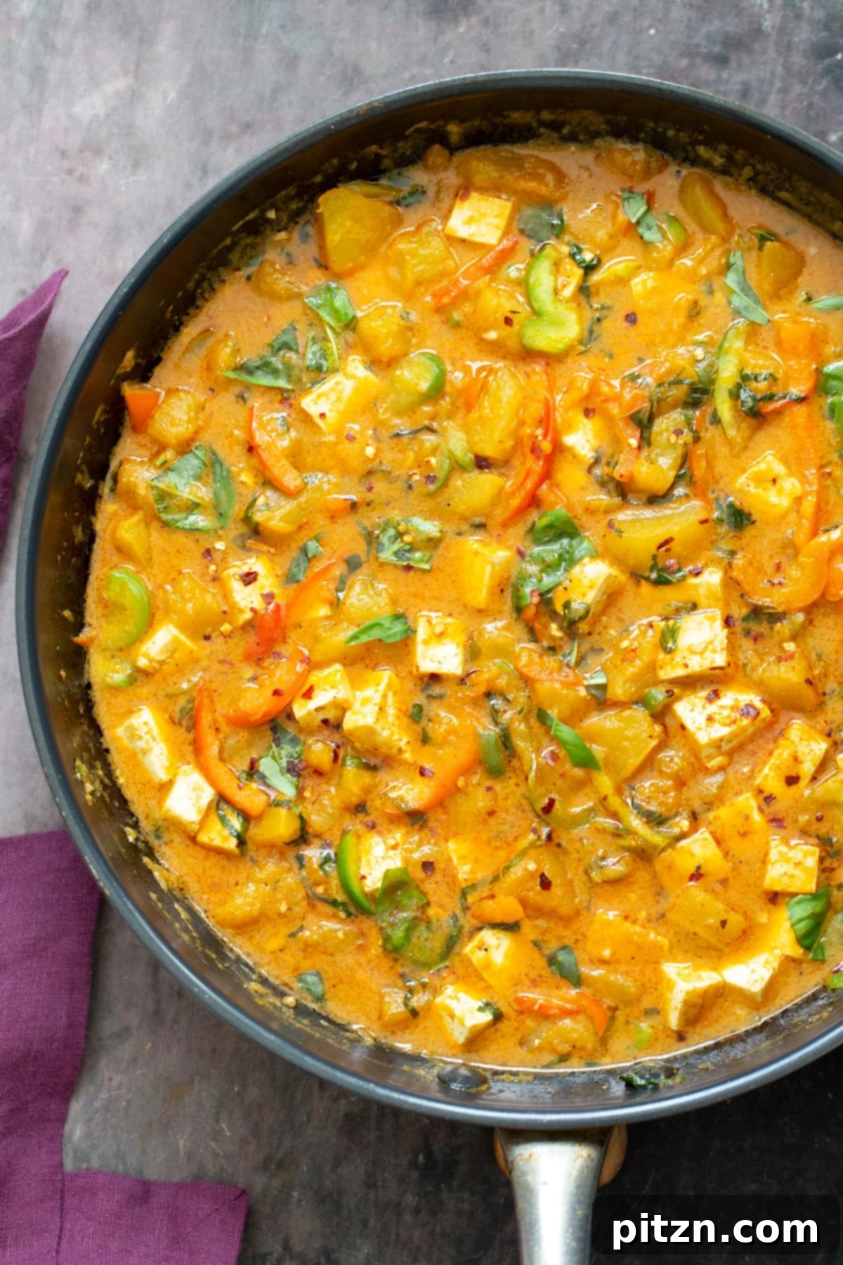A large black skillet brimming with freshly cooked Vegan Thai Pumpkin Curry, showcasing vibrant orange pumpkin cubes and green bell peppers in a creamy red sauce.