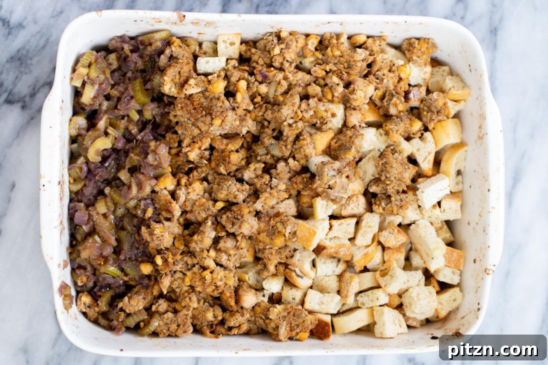 Various components for vegan stuffing—toasted bread, cooked vegetables, and chickpea sausage—being gently combined in a large white casserole dish.
