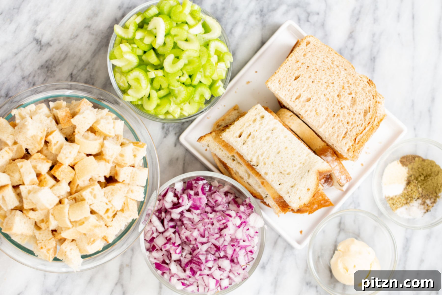 A selection of ingredients for vegan stuffing, including bread, celery, onions, and herbs, laid out on a marble countertop.
