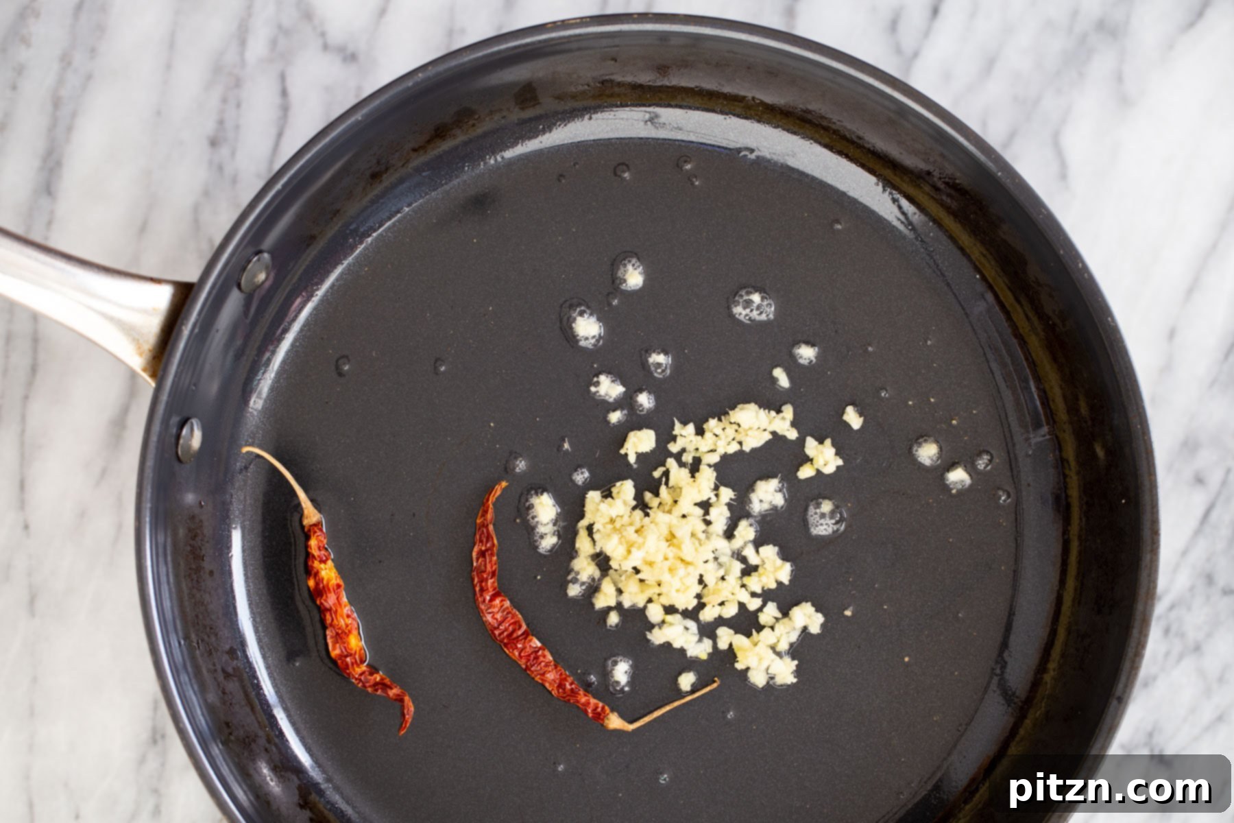Minced garlic and dried red chilies sizzling in hot oil in a black sautéing pan, infusing aromatic flavors.