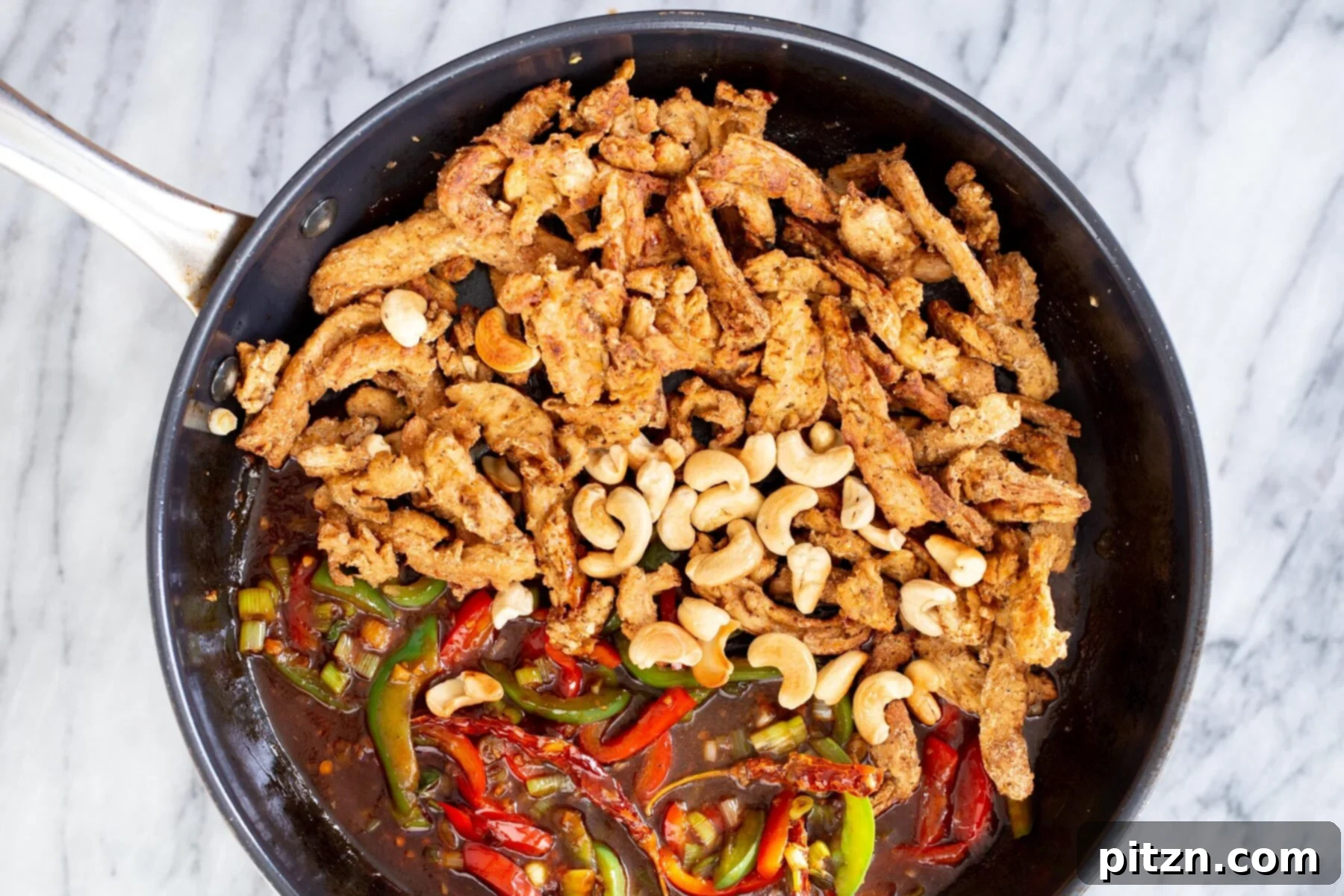 Crispy soycurls and toasted cashew nuts being added to the stir-fried vegetables and sweet and spicy sauce in a pan.