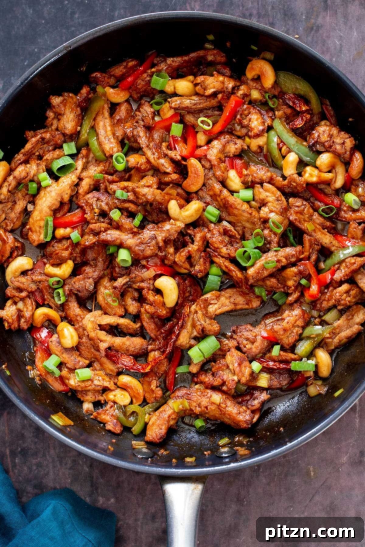 Chopped spring onions sprinkled over sizzling soycurls, cashews, and bell peppers in a sautéing pan, highlighting the preparation process.