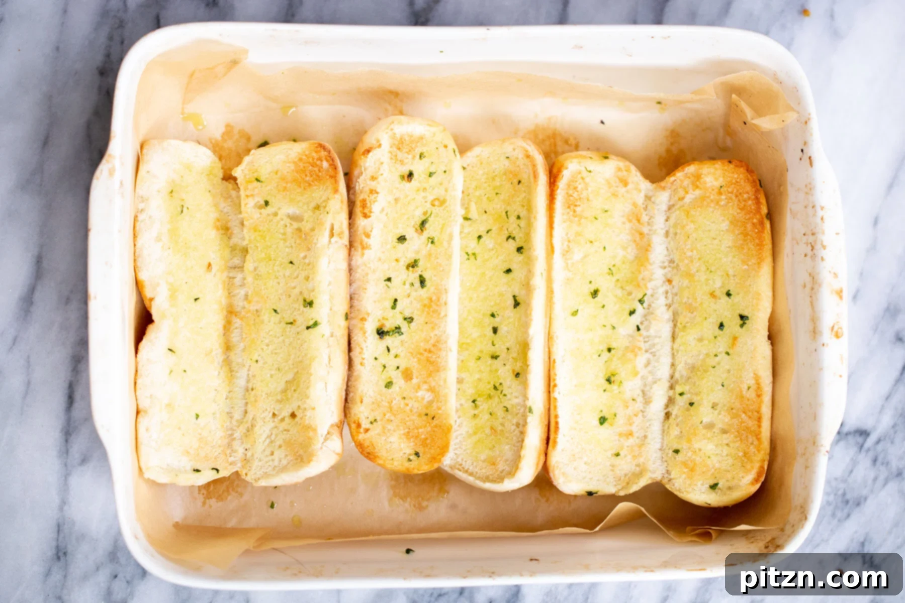 Sliced Italian baguette buns brushed with vegan butter, placed in a white casserole dish for toasting