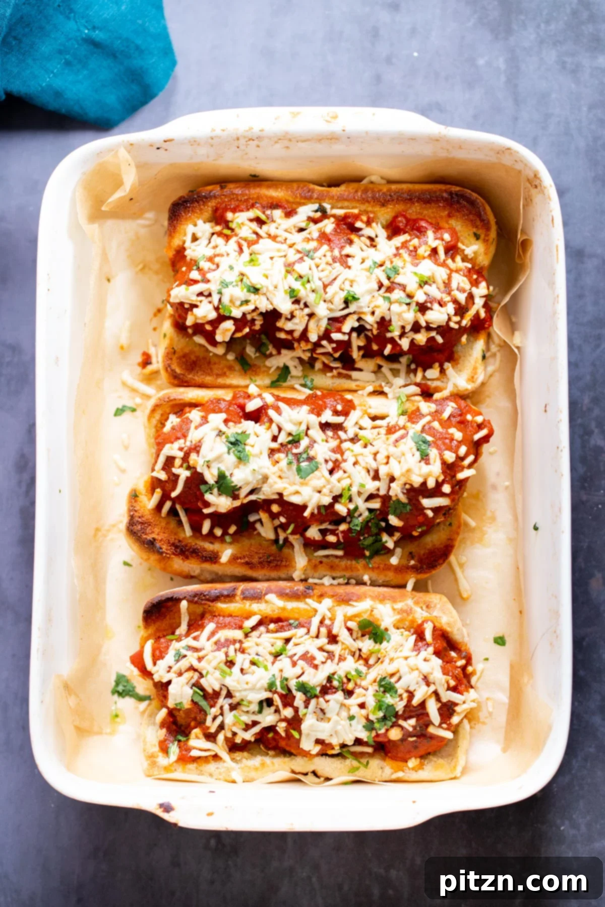 A close-up shot of several vegan meatball subs in a baking dish, adorned with succulent lentil meatballs, melted vegan cheese, and fresh green herbs