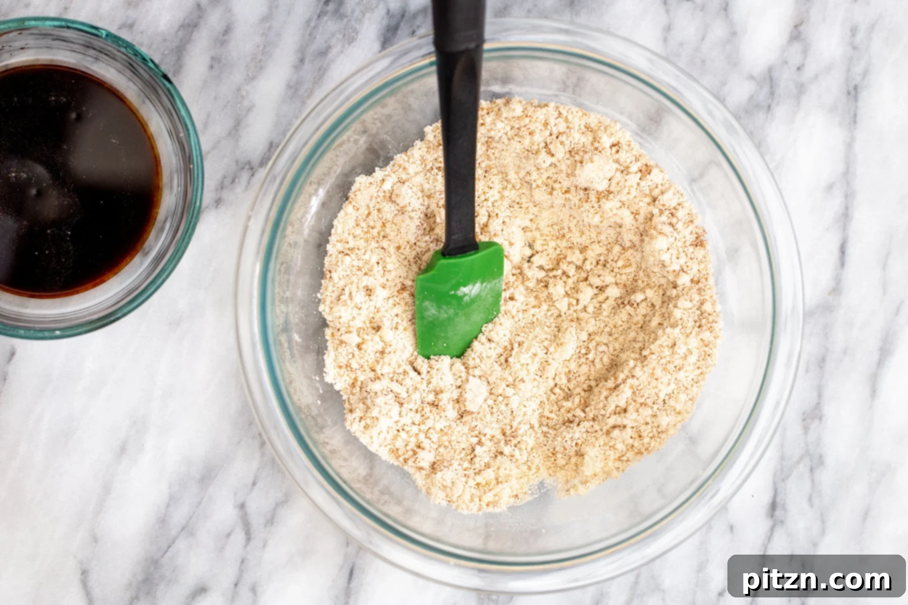 A pristine glass bowl containing well-mixed dry ingredients sits on an elegant marble countertop, anticipating the wet components for vegan ginger cookies.