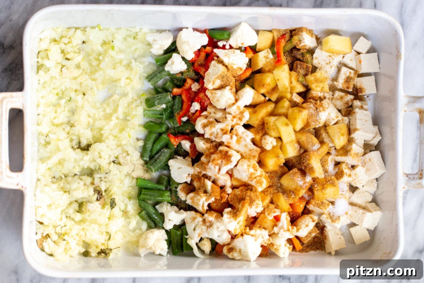 Ground spices being sprinkled evenly over a mix of fresh vegetables and tofu cubes in a white casserole dish.