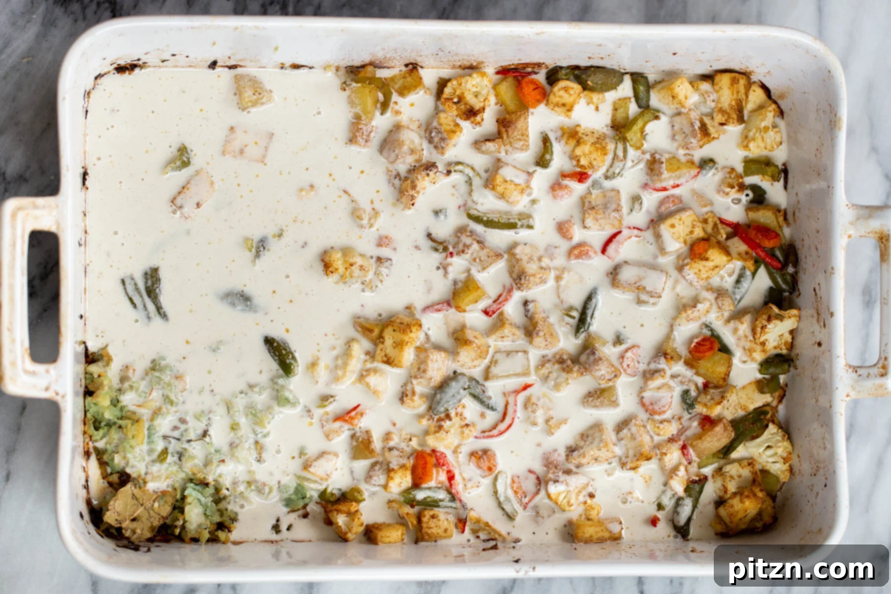 Rich, creamy cashew sauce being poured over the roasted vegetables and tofu in a white casserole dish, ready for the final bake.