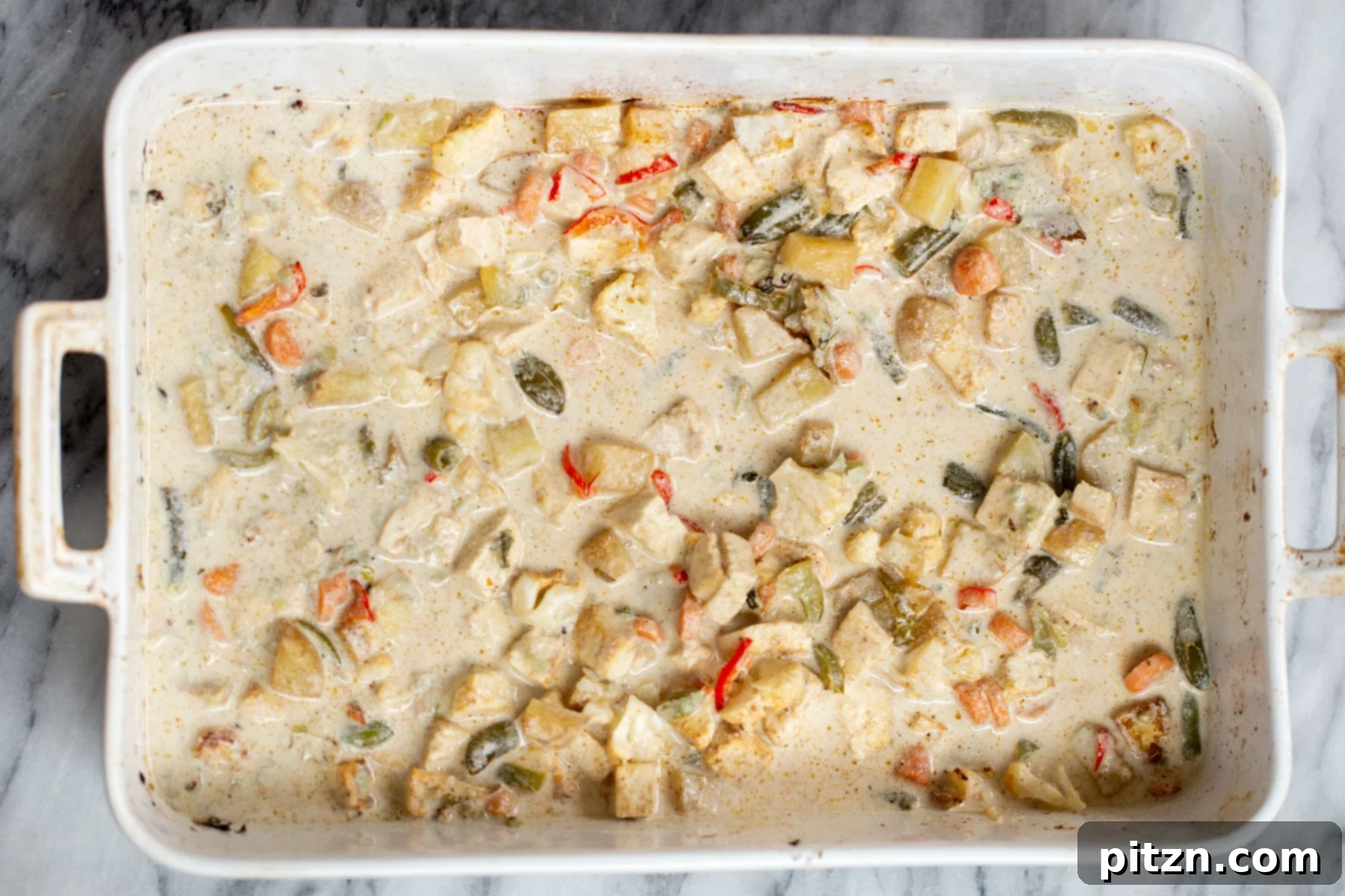 A close-up of creamy cashew sauce being generously poured on top of the roasted vegetables and tofu in a casserole dish, showcasing its smooth texture.