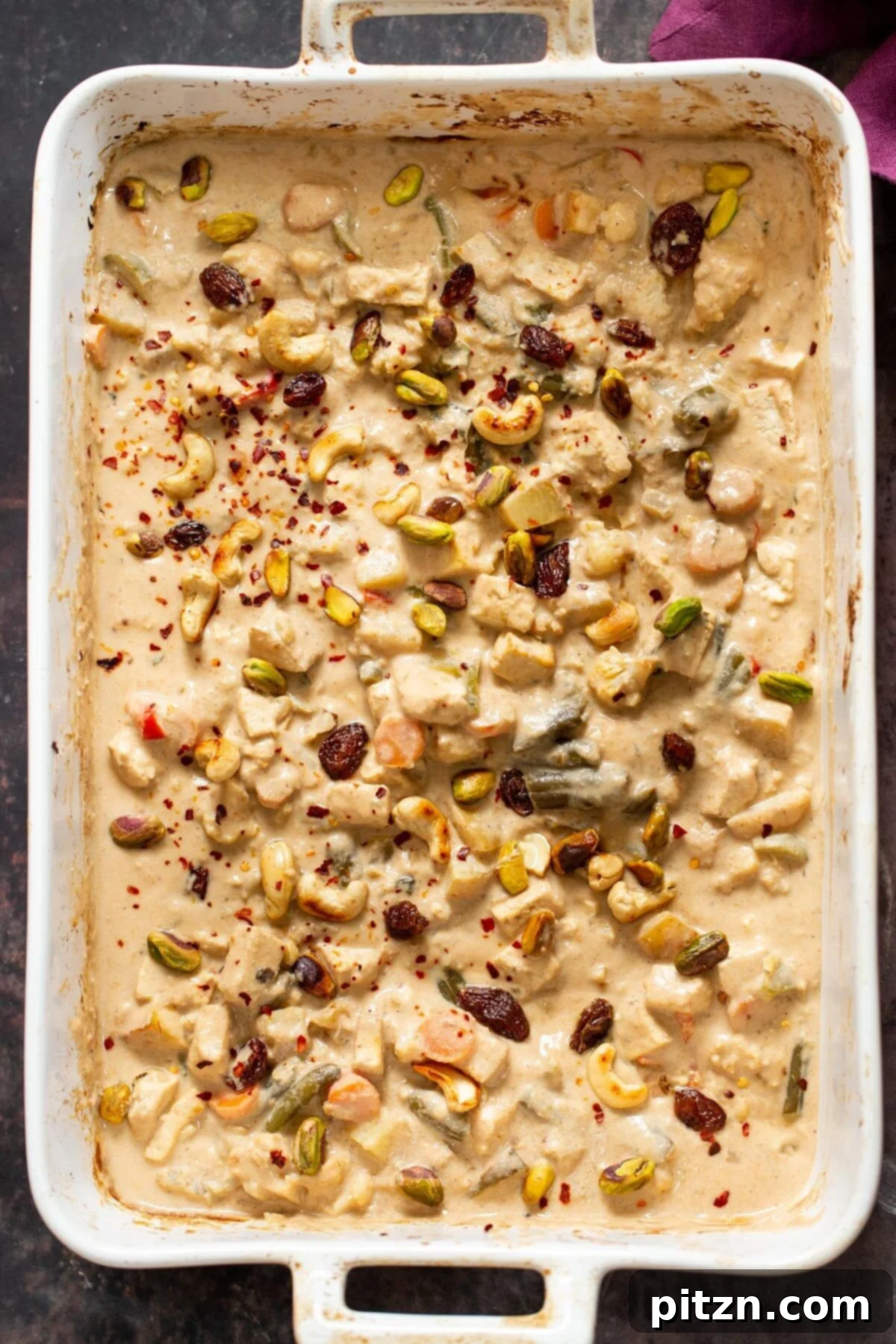 A close-up of creamy Baked Vegan Navratan Korma, being sprinkled with chili flakes and chopped nuts, highlighting its rich texture and vibrant garnish.