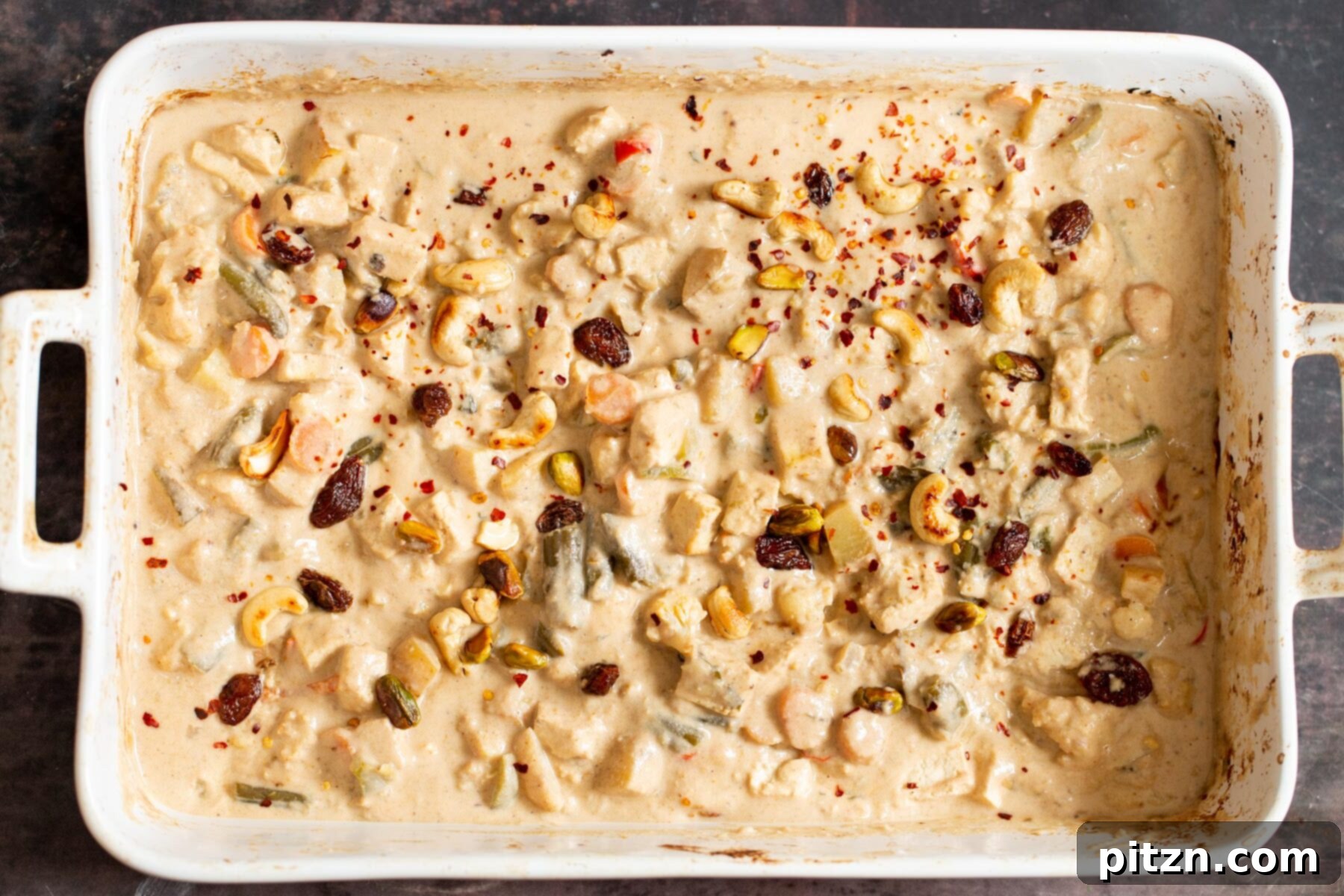 A white casserole dish showcasing creamy Indian Navratan Korma, generously sprinkled with toasted nuts and red chili flakes for a beautiful and delicious finish.