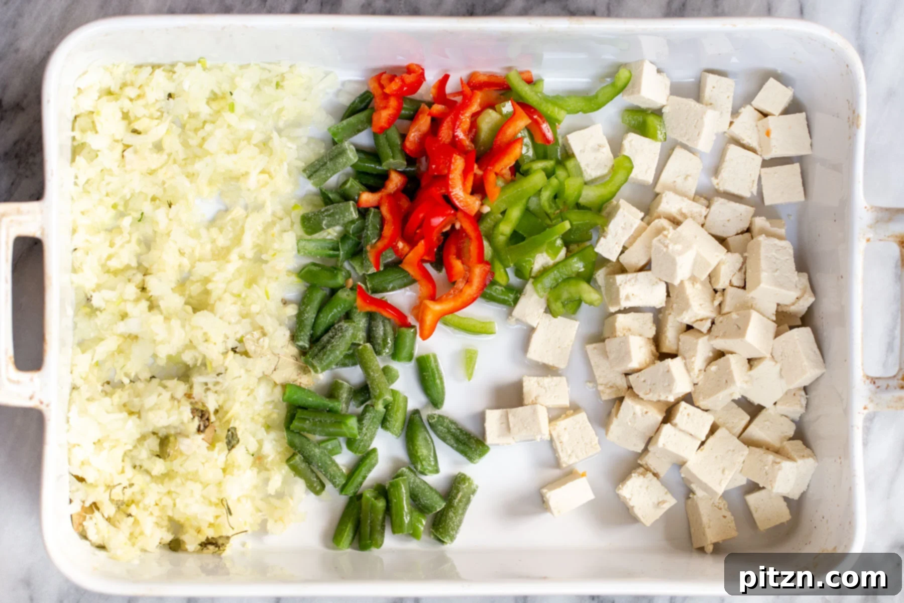 A large white casserole dish neatly arranged with finely chopped onions, ginger, and garlic on one side, and mixed vegetables and tofu cubes on the other, ready for baking.