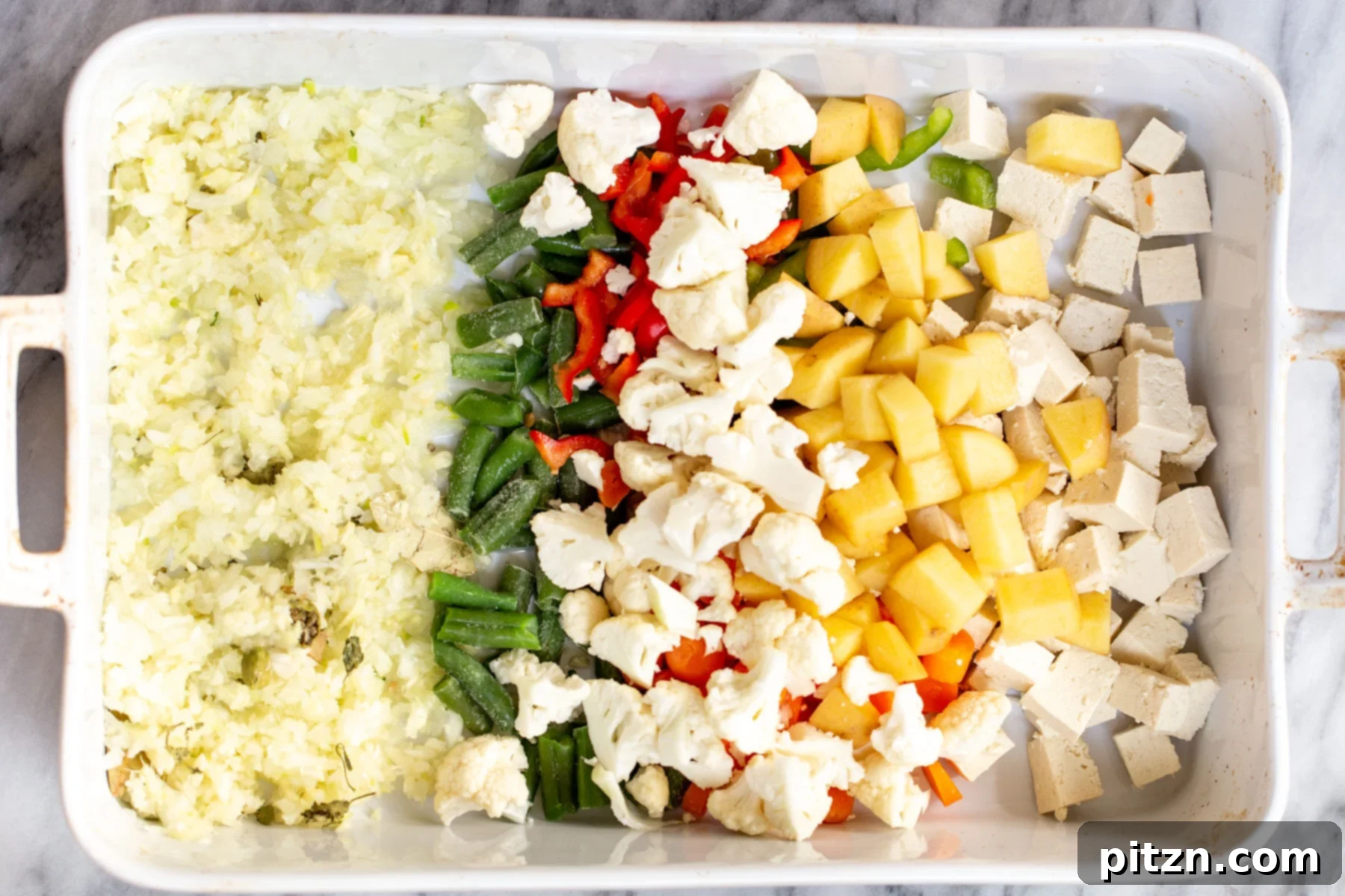Close-up of tofu cubes, mixed vegetables, and minced ginger and garlic in a white casserole dish, seasoned with ground spices.