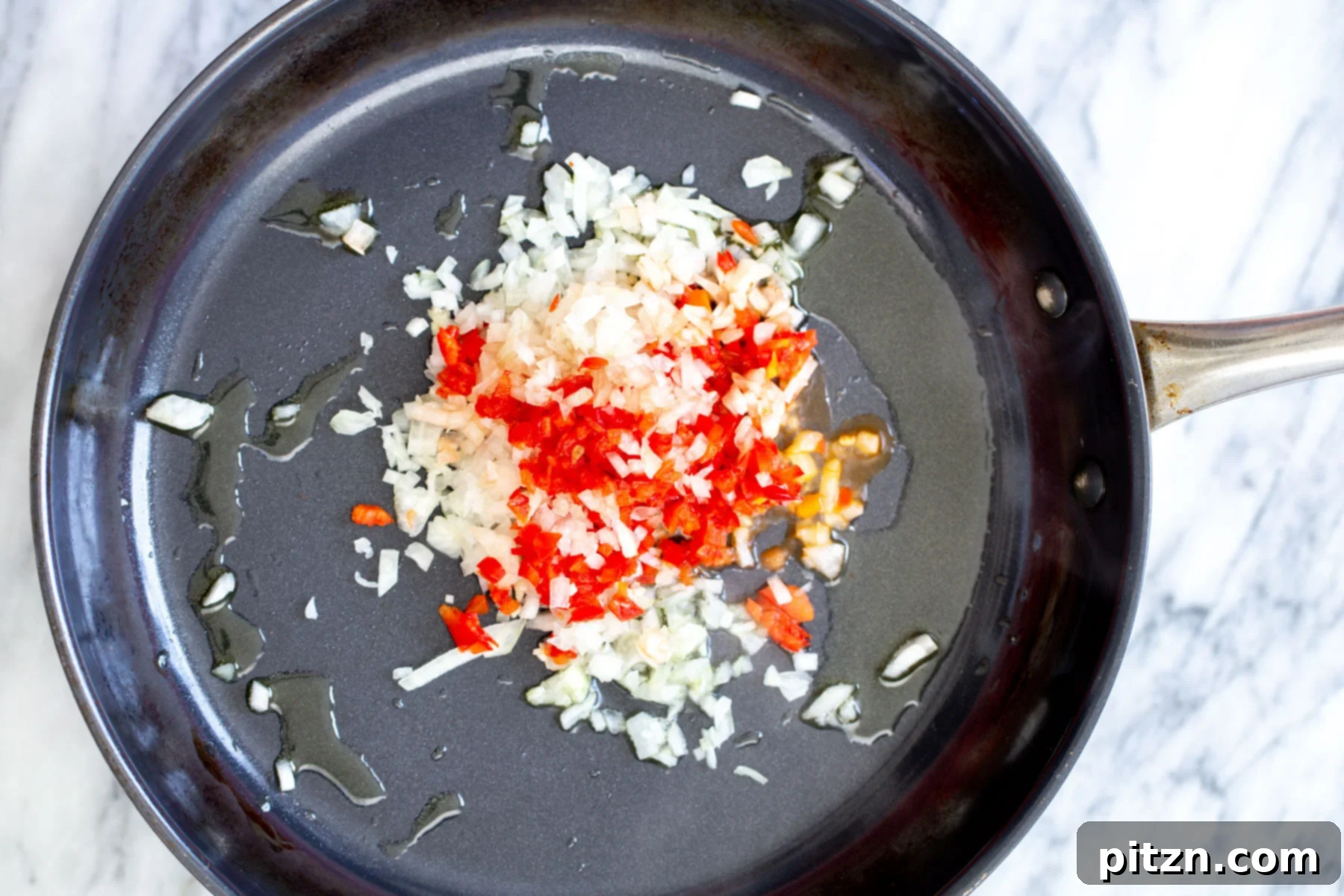 Minced red chili, garlic, and onion sautéing in a black pan, forming the aromatic base for the pasta sauce.