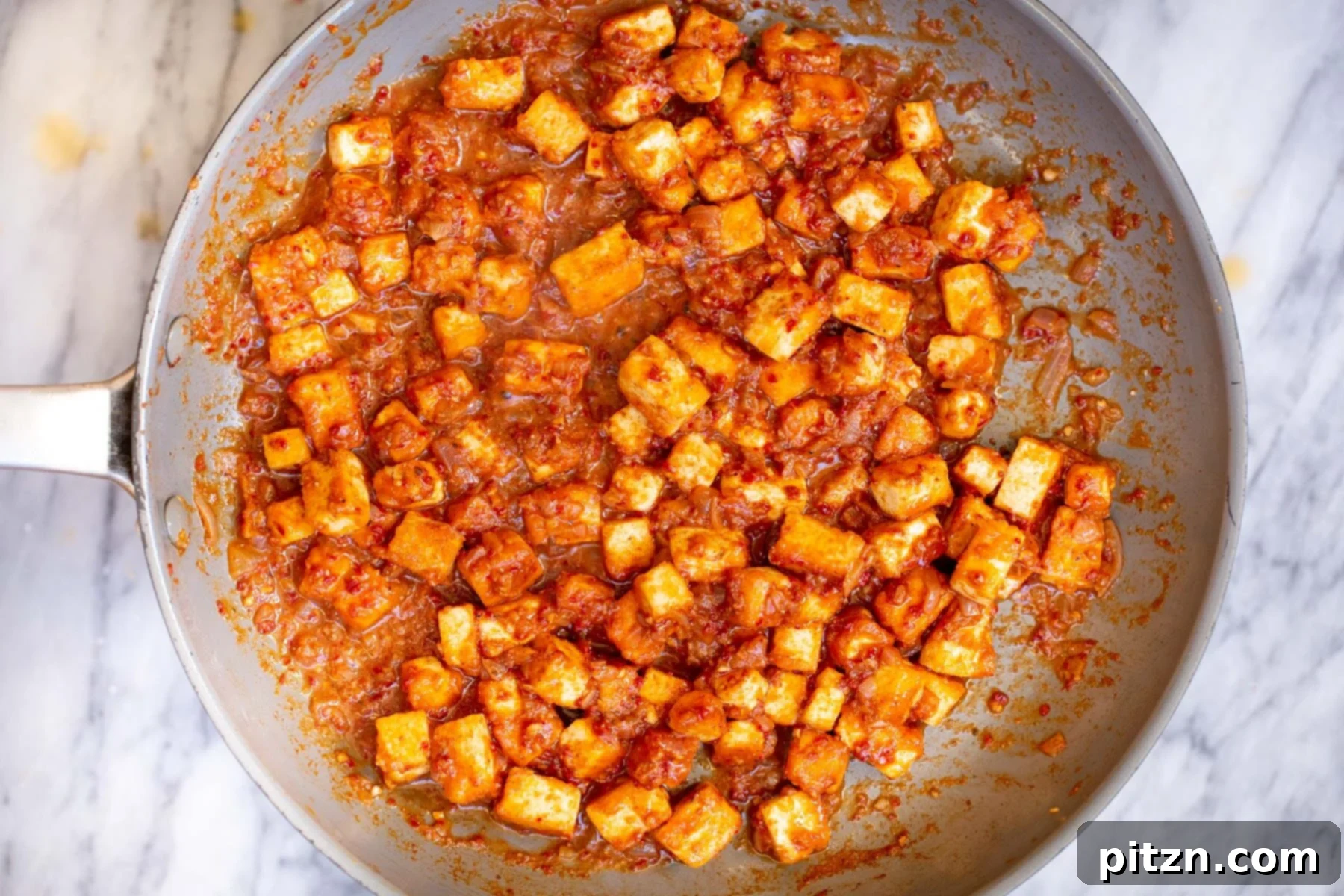A closeup of finished Tofu Vindaloo simmering gently in a sauteing pan on a marble countertop, rich and inviting.