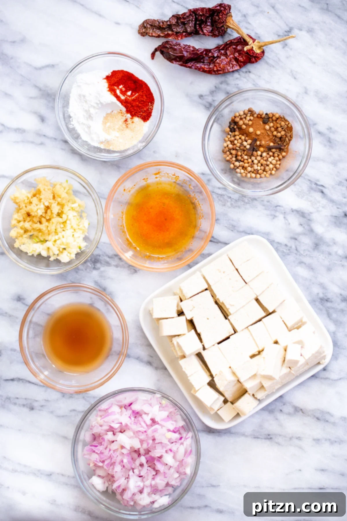A collection of fresh ingredients laid out on a marble countertop, including various spices, chilies, ginger, garlic, and vinegar, ready for making Tofu Vindaloo.