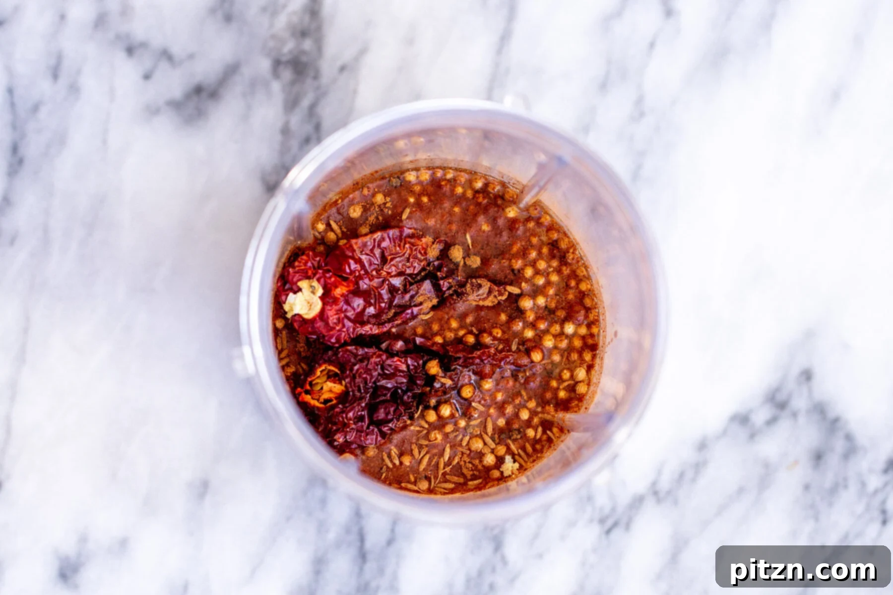 Ingredients for vindaloo chili paste, including whole spices and soaked chilies, arranged in small bowls on a marble countertop.