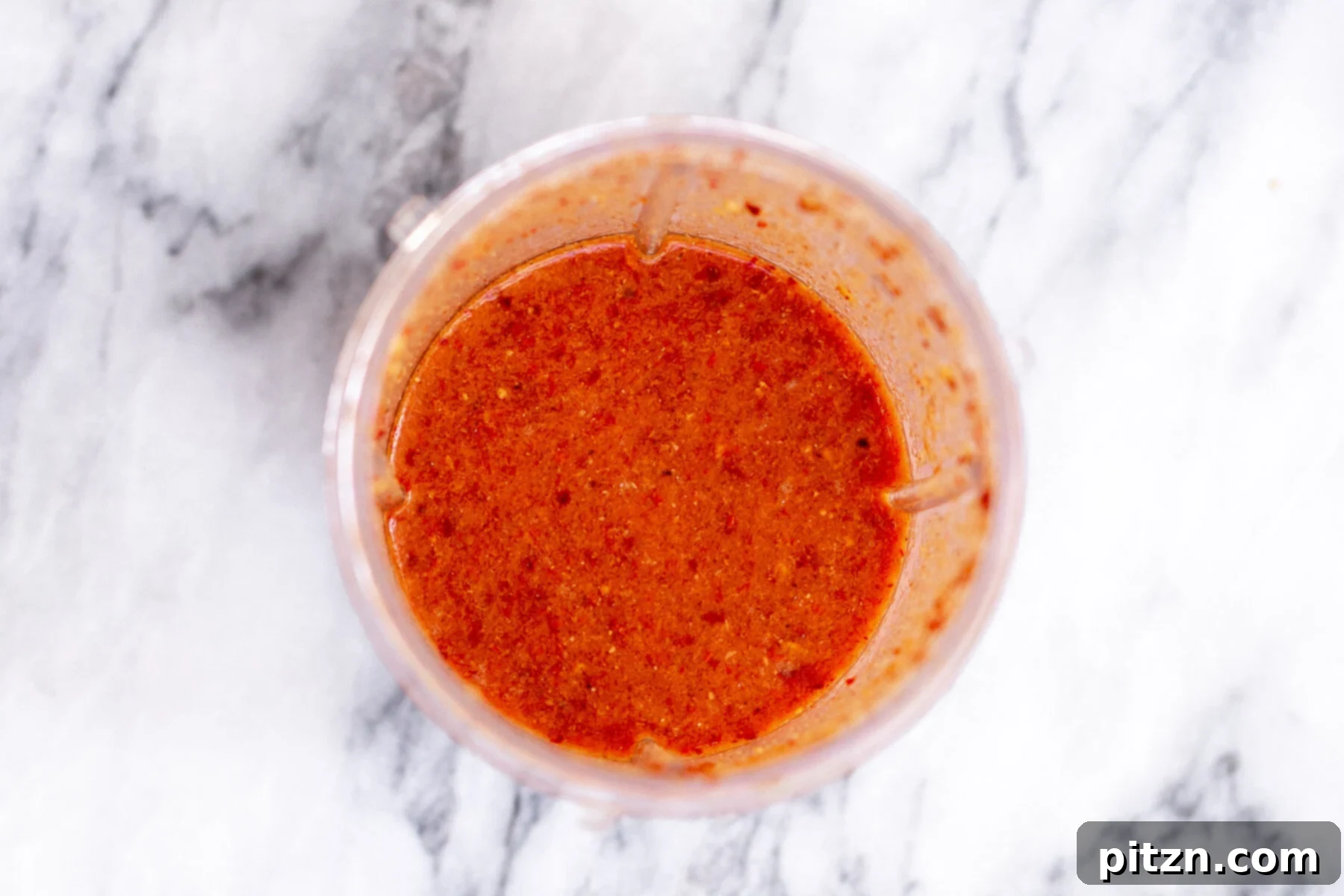 A small glass container holding the freshly blended, rich red vindaloo chili paste on a marble countertop.