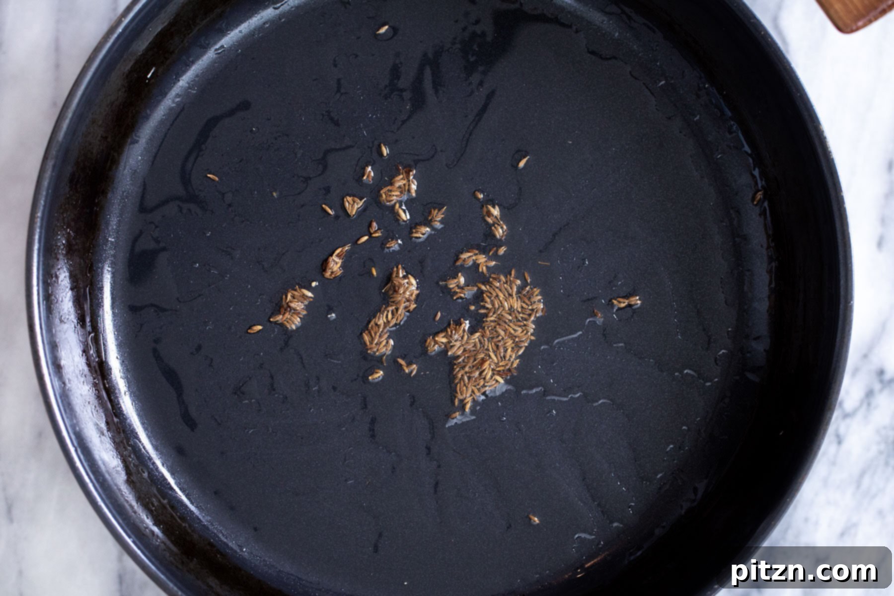 Cumin seeds sizzling in hot oil in a black sautéing pan, forming the aromatic base of the curry.