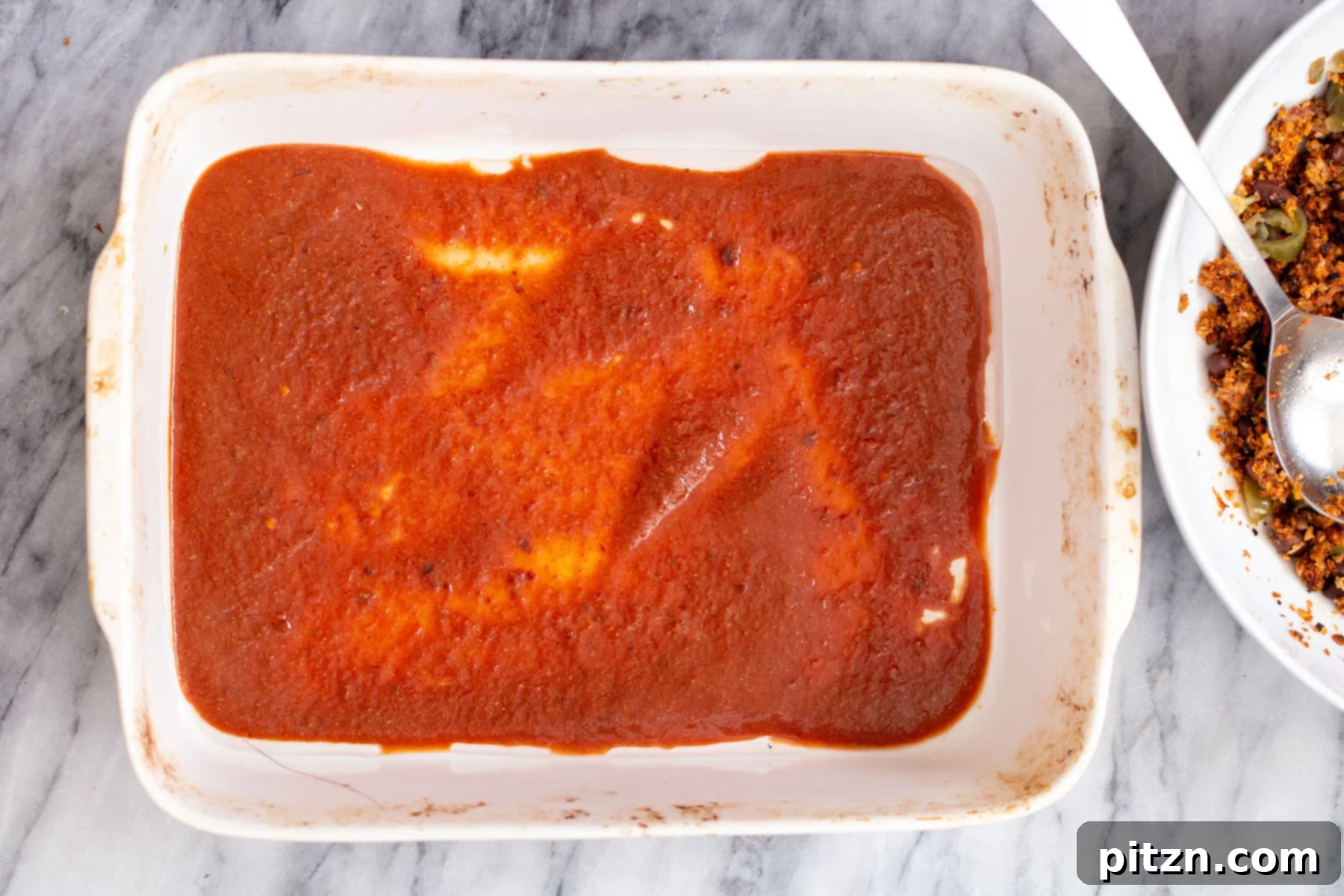 A layer of vibrant red vegan enchilada sauce being spread at the bottom of a casserole dish before adding the filled tortillas.