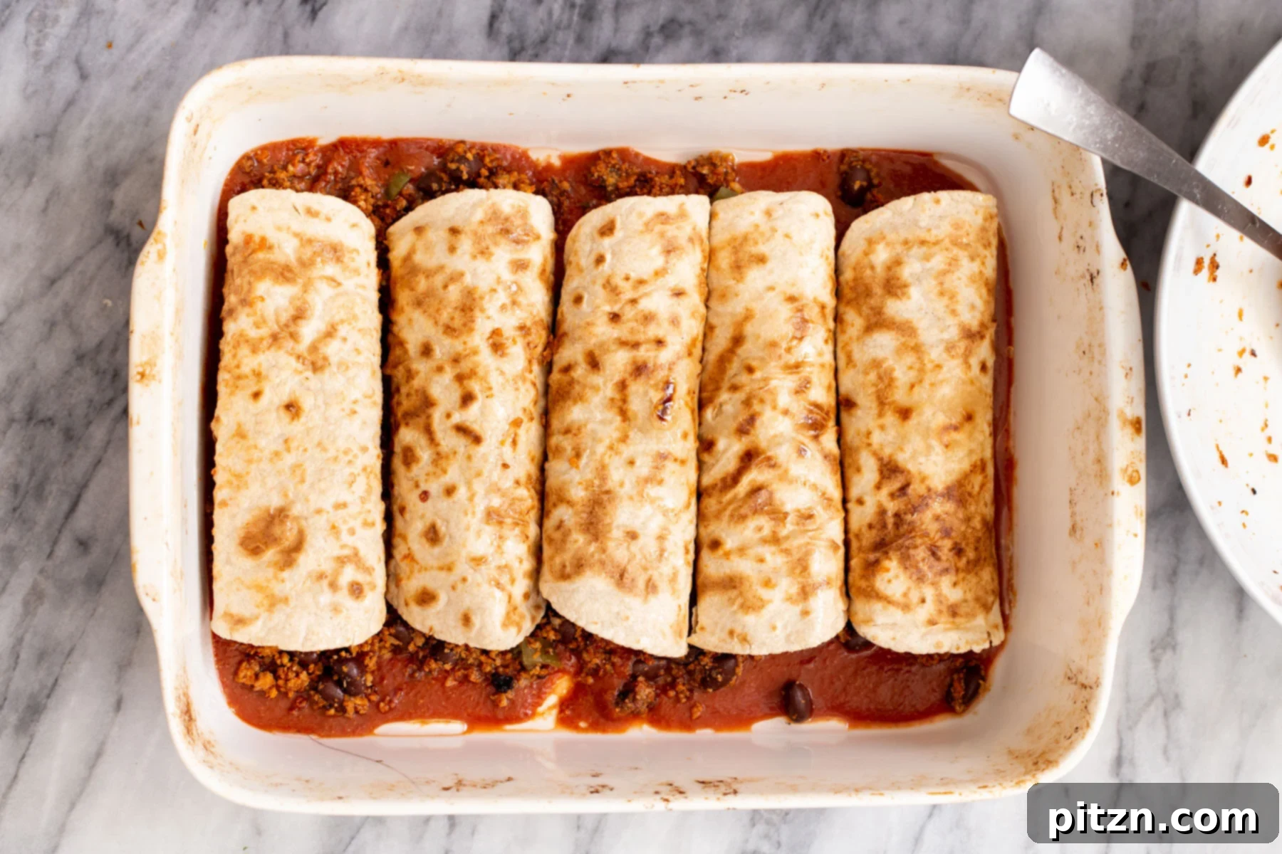 A casserole dish filled with rolled enchiladas, generously covered in a rich red tomato sauce, ready for the final bake.