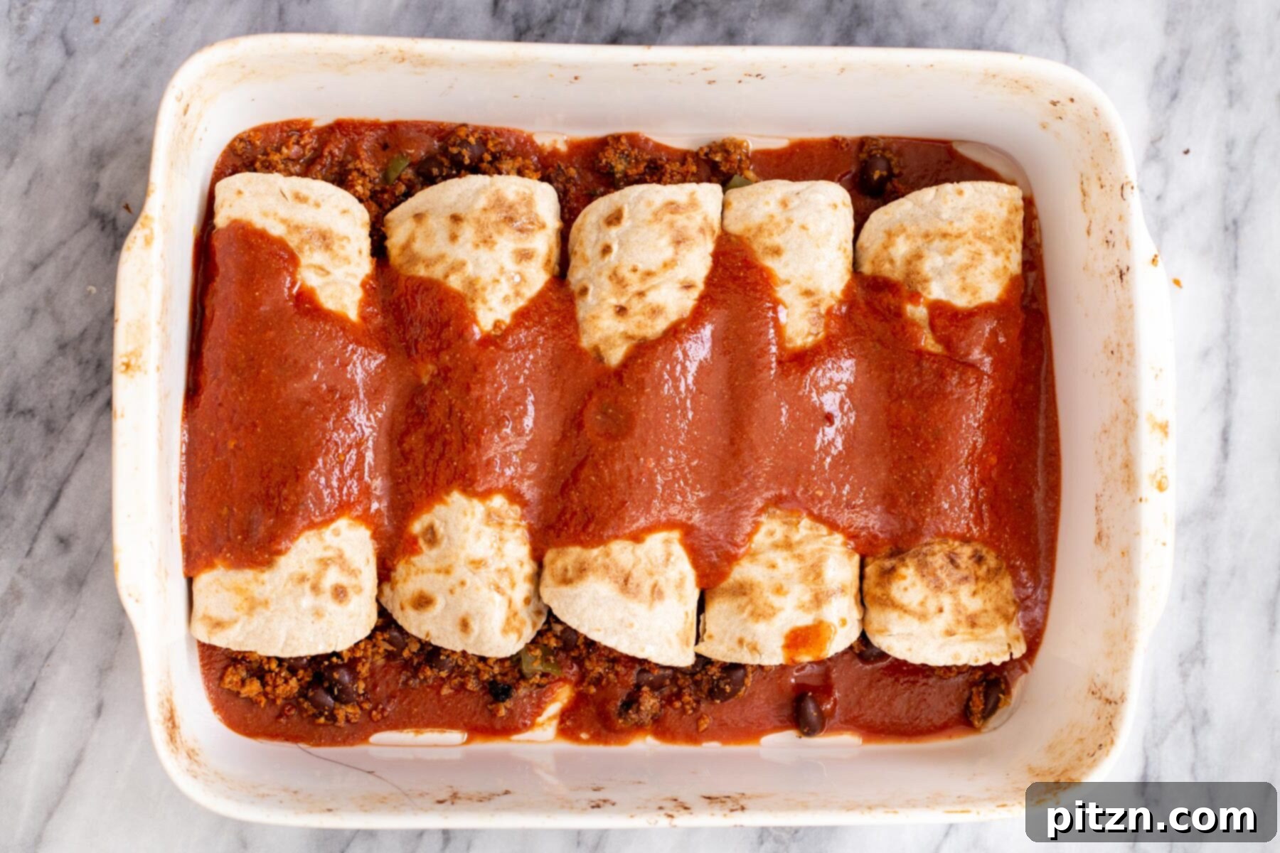 A casserole dish brimming with vegan taco meat stuffed enchiladas, beautifully covered with a rich red salsa, freshly baked.