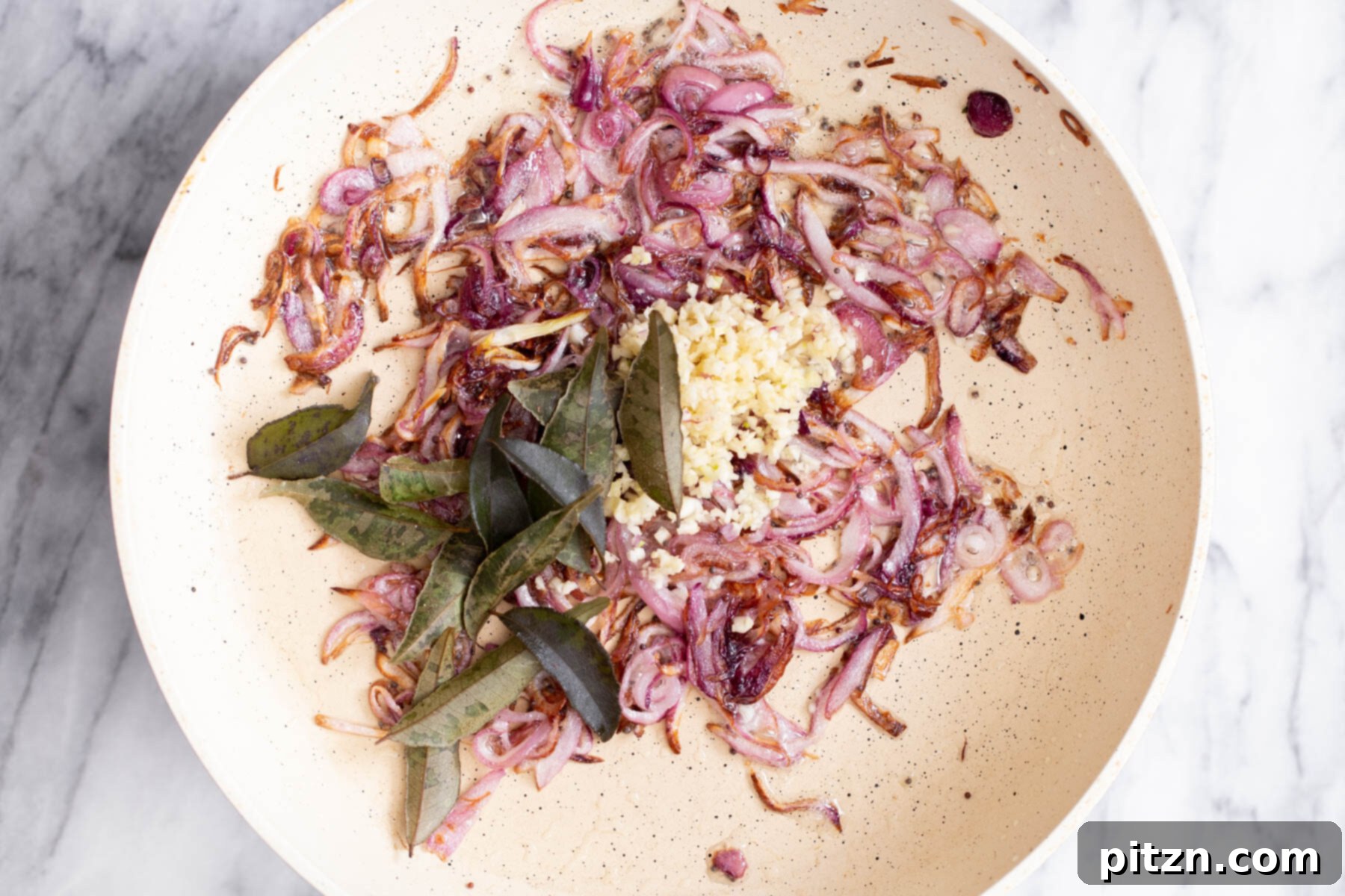 Minced garlic cloves and vibrant green curry leaves being combined with the browned onions in the hot frying pan.