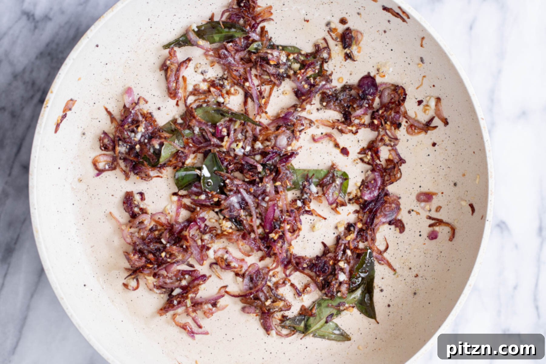 A fragrant medley of fried onion, garlic, and curry leaves simmering in a sautéing pan, forming the aromatic base of the dish.