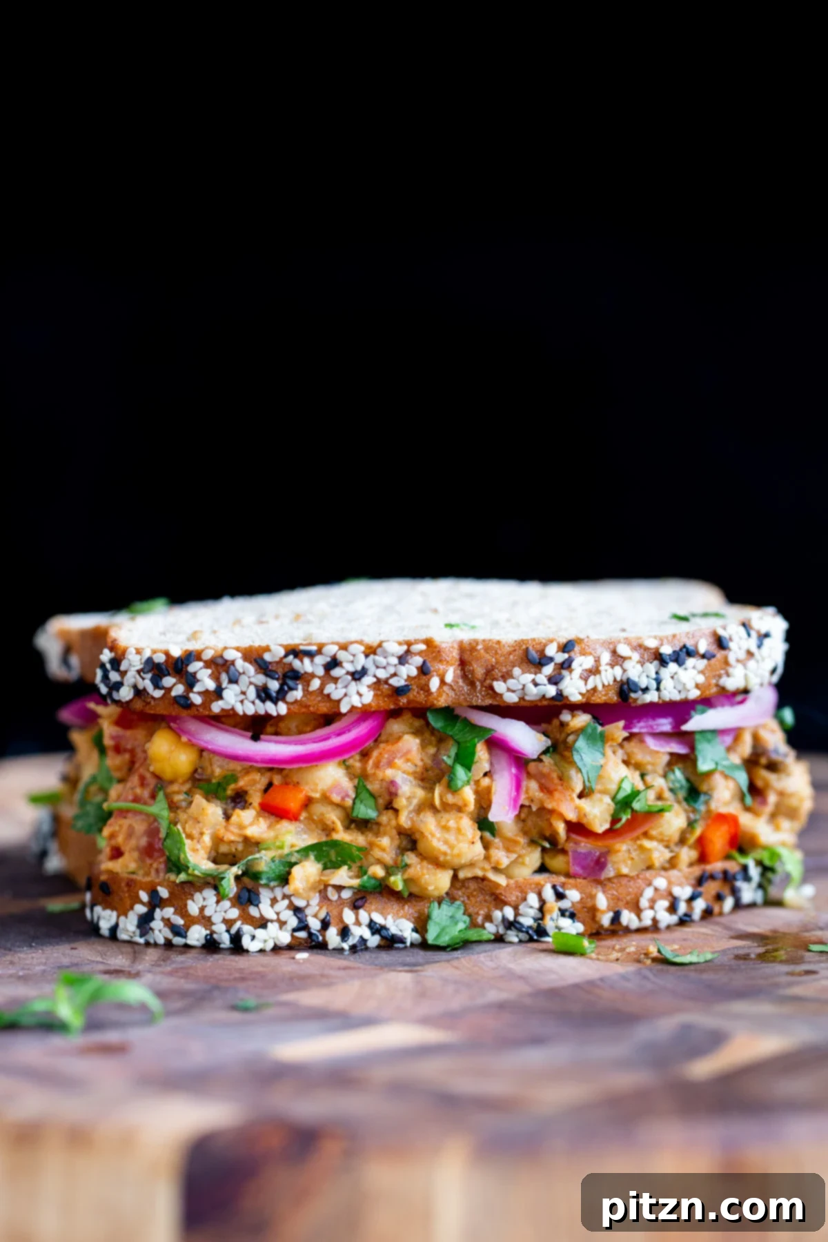 Delicious vegan curried chickpea salad sandwich on a wooden cutting board, garnished with fresh cilantro and pickled red onions