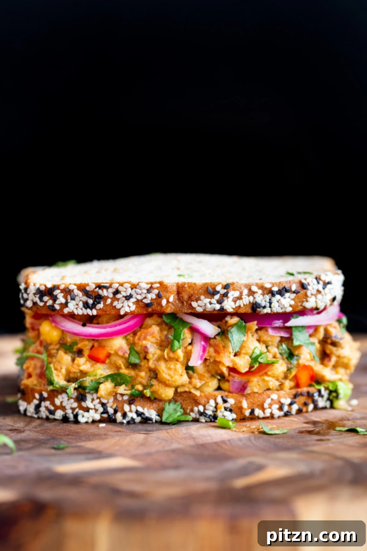 A finished vegan curried chickpea salad sandwich, perfectly cut and presented on a wooden chopping board with fresh herbs