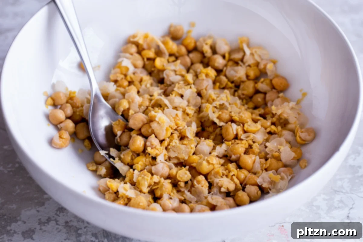 Drained chickpeas being mashed in a white mixing bowl with a fork, ready for seasoning