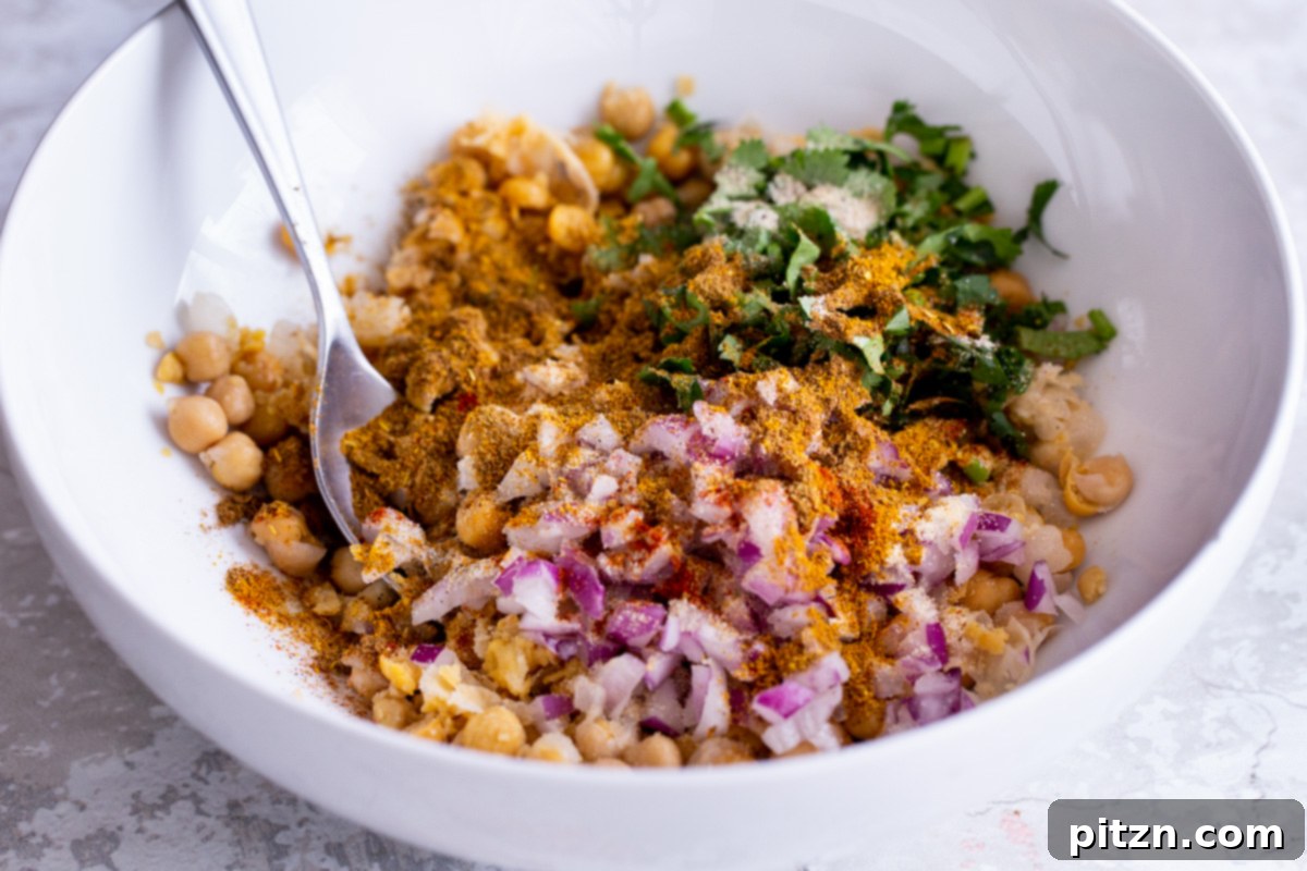 Spices, chopped red onions, and fresh herbs being added to mashed chickpeas in a white bowl for vegan curried chickpea salad