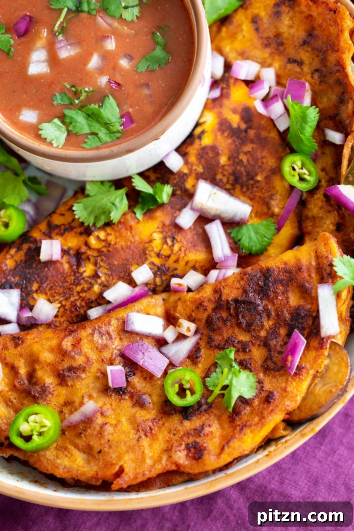 A vibrant plate of Vegan Birria Tacos, generously filled and served with a small bowl of rich, creamy Tikka Masala dipping sauce. Garnishings of fresh cilantro, diced red onions, and lime wedges complete this irresistible fusion dish.