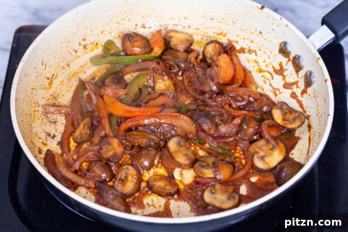 The vegan birria taco filling, a vibrant mixture of sautéed mushrooms, bell peppers, and onions, is simmering gently in a skillet, now enriched with the creamy Tikka Masala sauce. The vegetables are coated in the orange-hued gravy, ready for taco assembly.