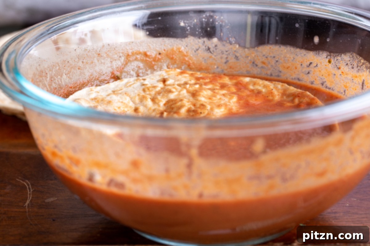 A corn tortilla is being expertly dipped into a small glass dish containing the rich orange Tikka Masala gravy, ensuring it is thoroughly coated before being placed onto the hot skillet.