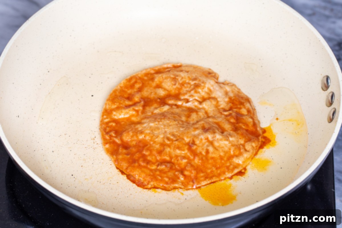 A sauce-coated tortilla rests on a hot skillet, with a scoop of the flavorful vegan birria filling and a sprinkle of shredded vegan cheese added to one half, ready to be folded and cooked until golden and crispy.