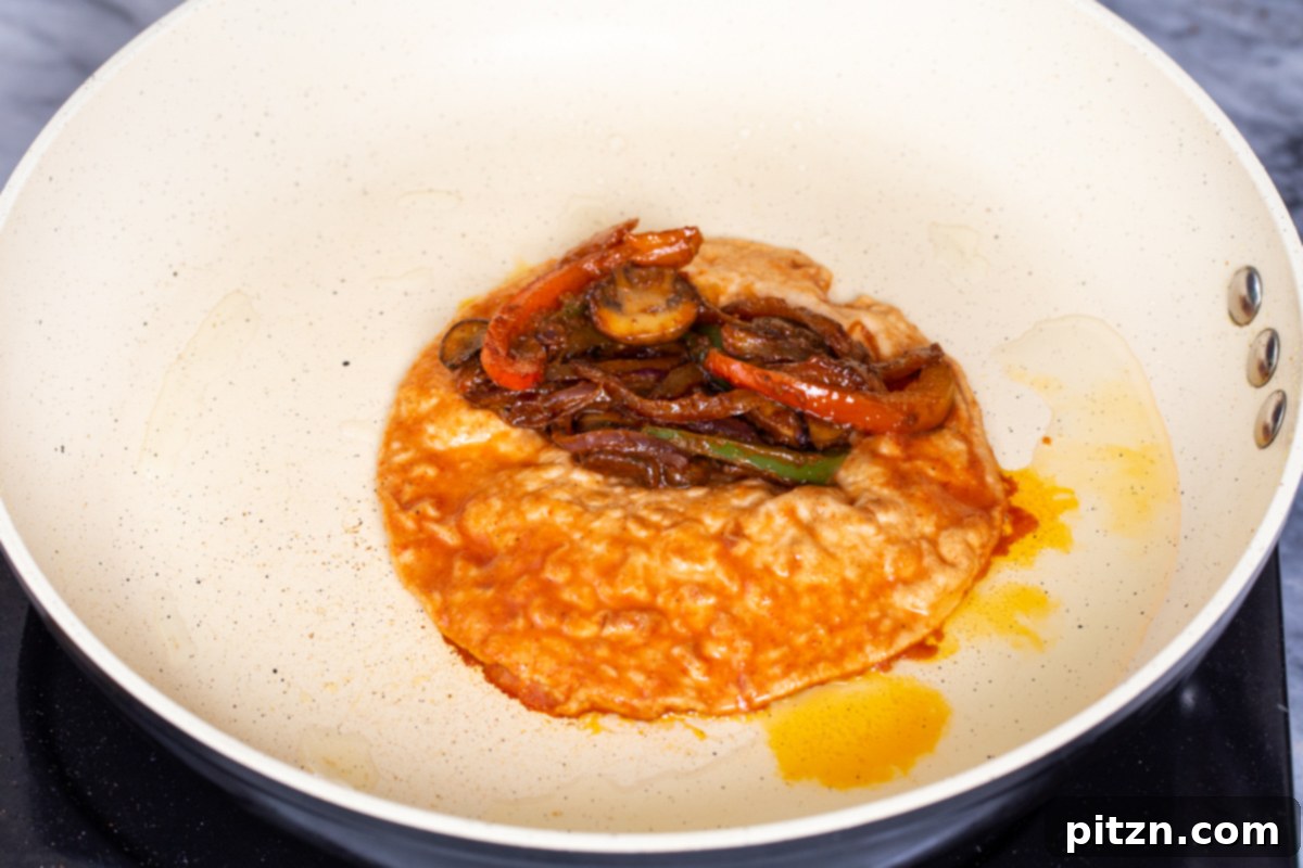 A golden-brown tortilla in a skillet, topped with roasted red bell pepper pieces and other elements of the vegan birria filling, indicating it's almost ready to be folded and pressed into a savory taco.