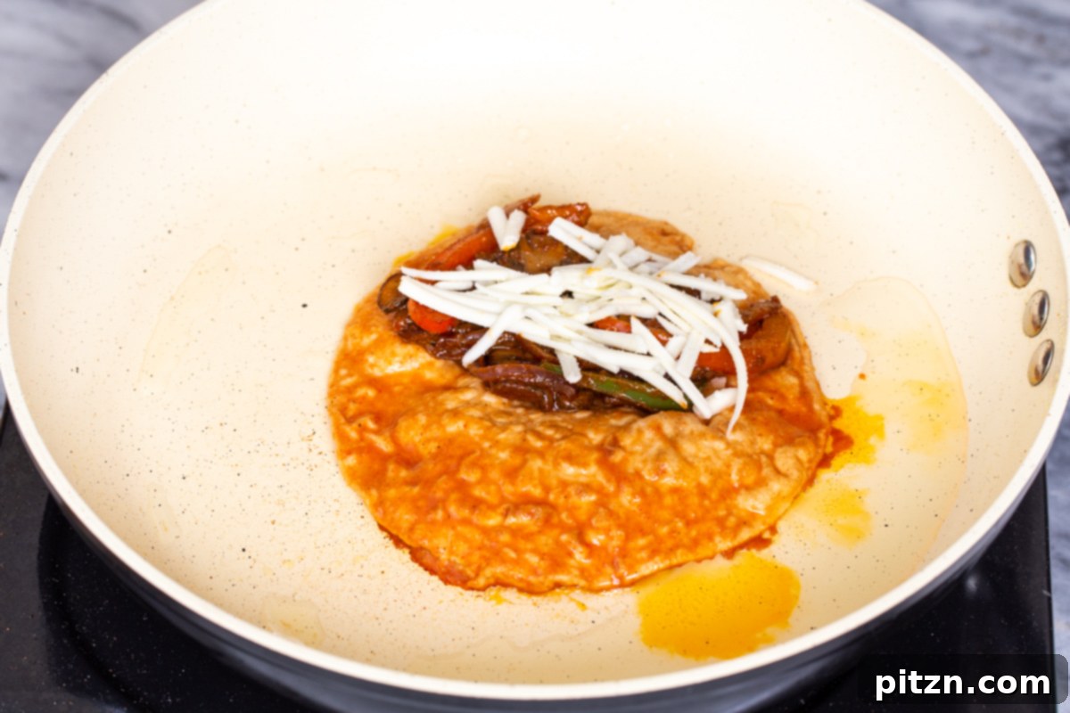 A half-folded tortilla in a skillet, filled with a mix of vibrant vegetables and generously topped with shredded vegan cheese. It's being pressed down to melt the cheese and crisp the tortilla, creating a delectable vegan birria taco.