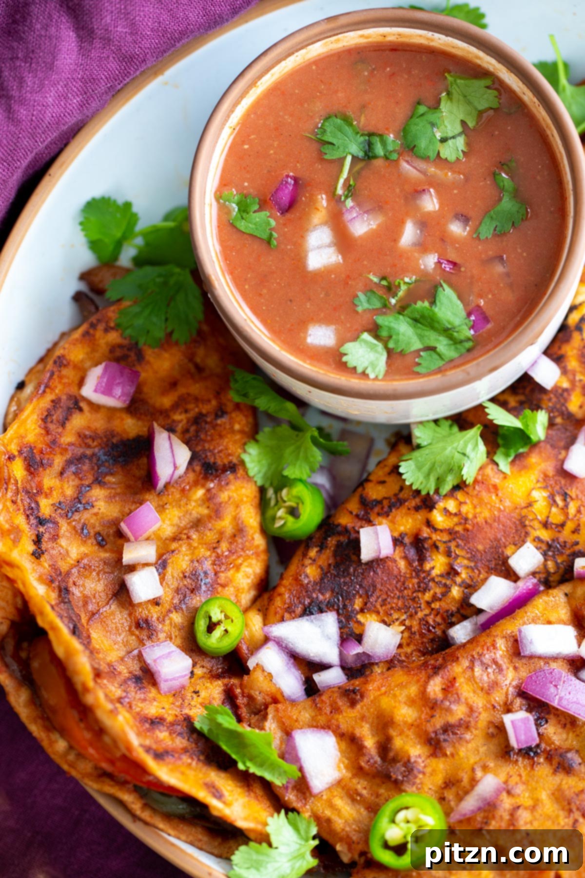 A beautifully presented plate of Vegan Birria Tacos, garnished with fresh cilantro and diced red onions, is accompanied by a small bowl of rich, orange Tikka Masala dipping sauce. Lime wedges add a pop of color and freshness to this inviting fusion meal.