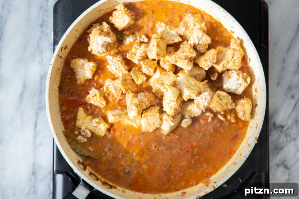 Crispy baked tofu chunks being gently introduced into the simmering Indian gravy in a frying pan.