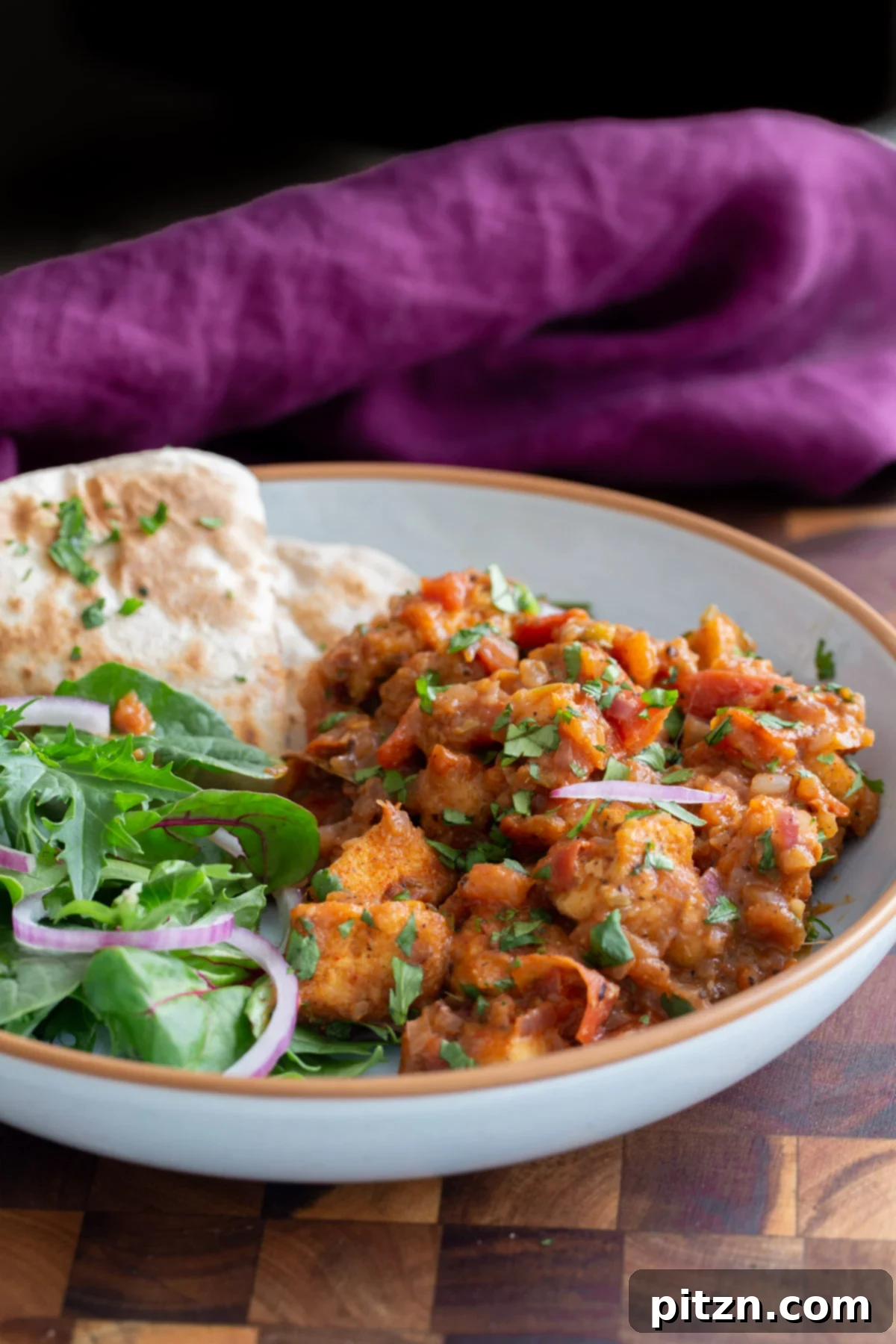 A captivating side view of a serving of vegan Khada Masala Murg curry, beautifully presented with fresh greens and warm flatbread, highlighting its rich texture.