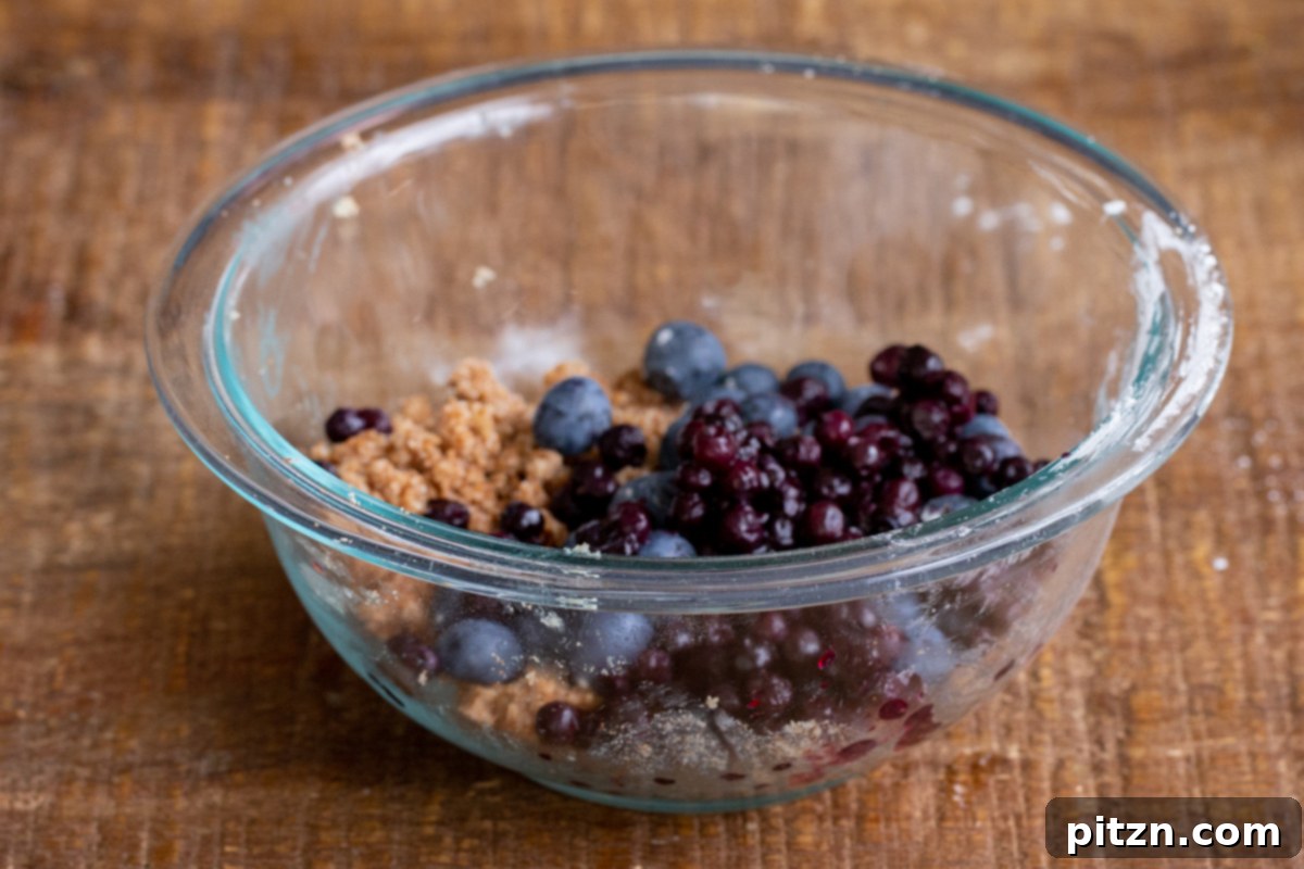 Gluten-Free Vegan Blueberry Muffin Cookies 11 Fresh and frozen blueberries being added to the streusel dough in a clear glass bowl, preparing for the topping of Vegan Blueberry Muffin Cookies.
