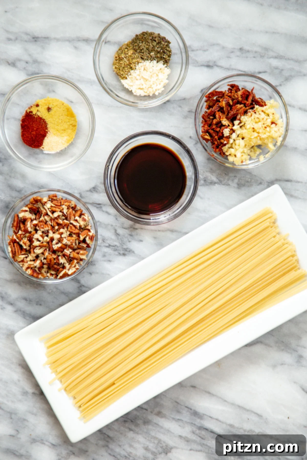 A flat lay of all the essential ingredients arranged for making vegan chili garlic oil pasta, showcasing fresh garlic, sun-dried tomatoes, spices, pasta, and pecans.