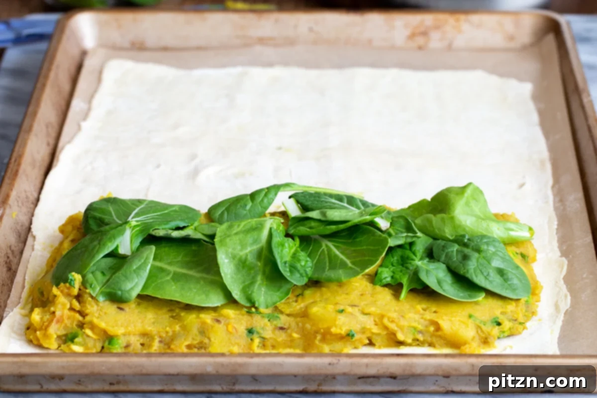 Fresh spinach leaves being added as a final layer over the potato filling during the assembly of the vegan samosa pie.